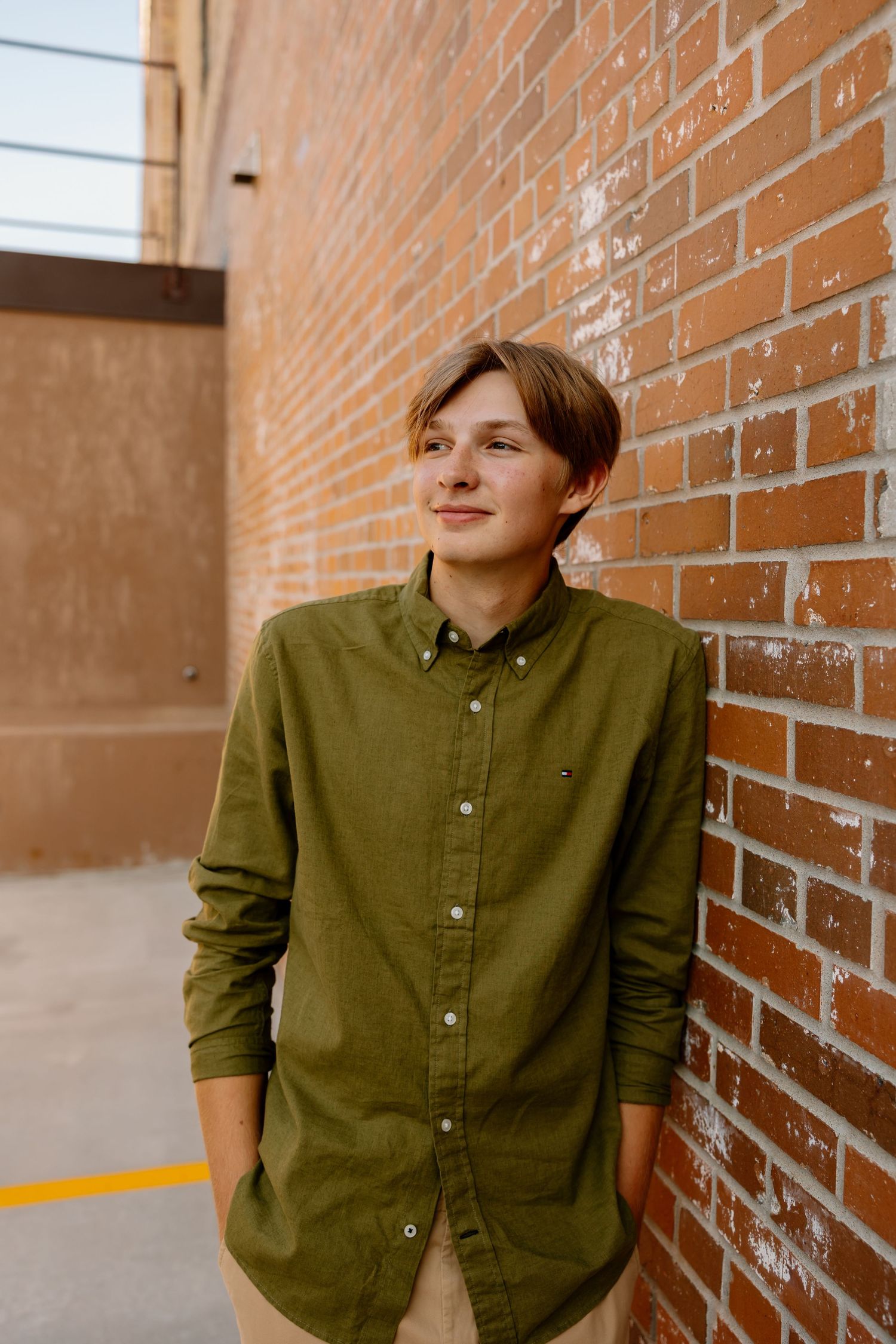 A person in an olive green button-down shirt poses against a brick wall in an urban setting.