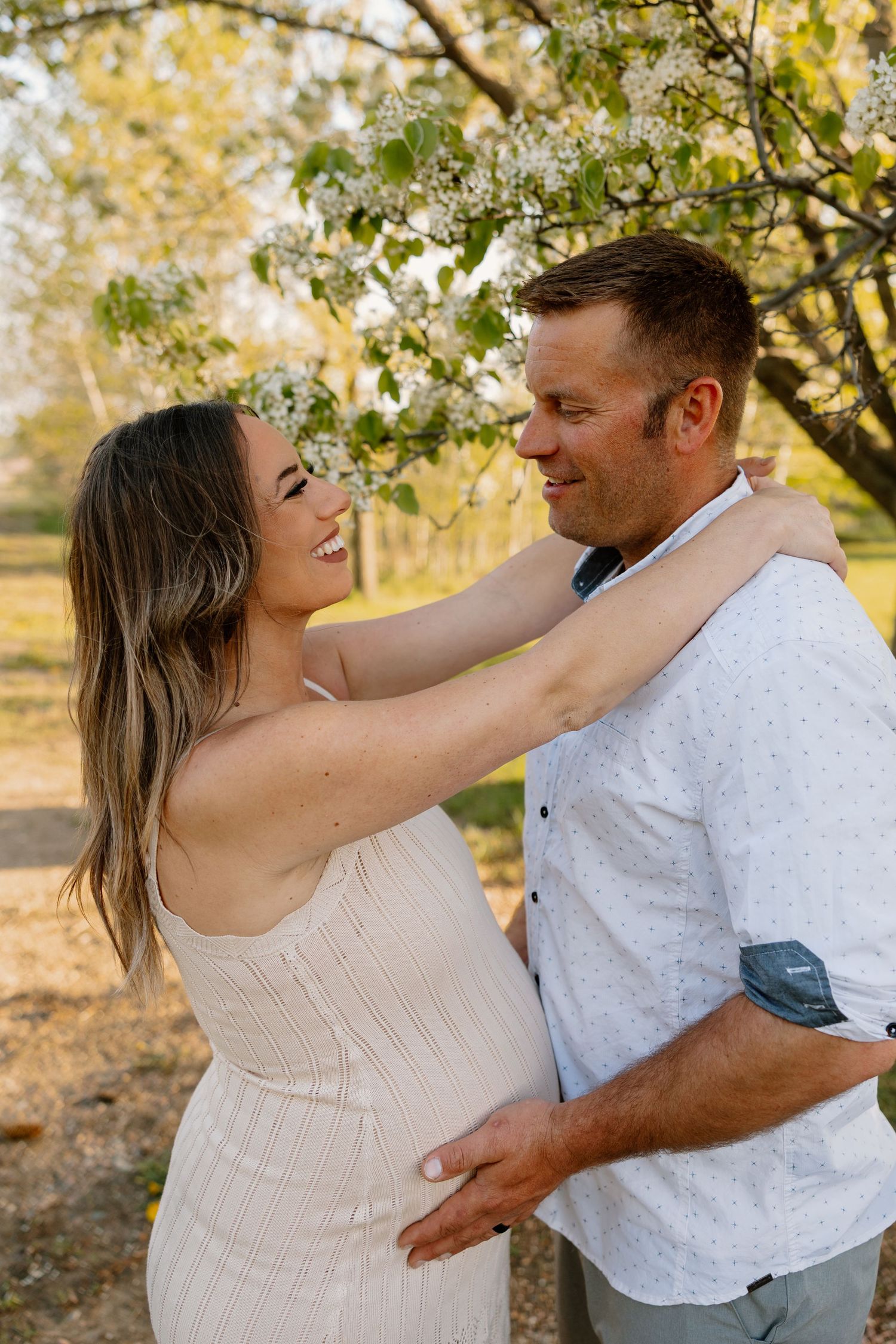 Couple embraces lovingly under autumn tree branches during golden hour sunset in soft neutral attire.
