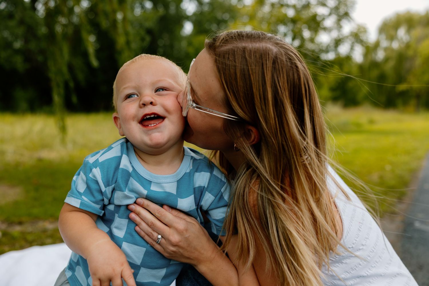 A joyful laughing baby in a blue outfit receives a loving kiss outdoors on a sunny day.