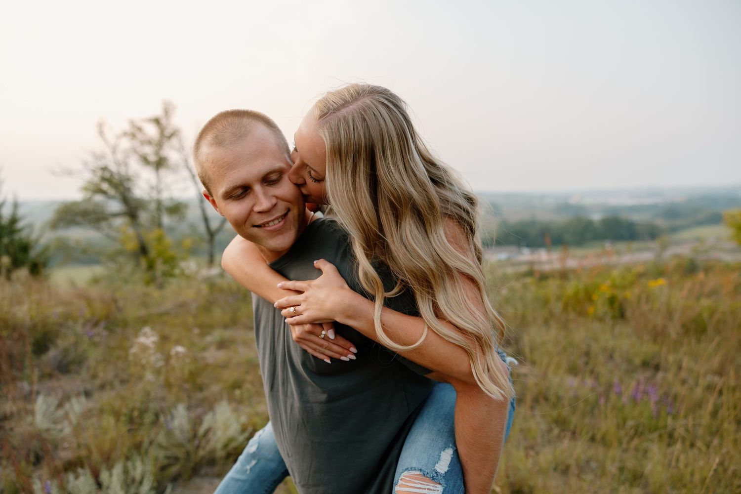 A playful couple sharing a piggyback ride and laughing together in a scenic outdoor setting during golden hour. sioux falls photographer ril