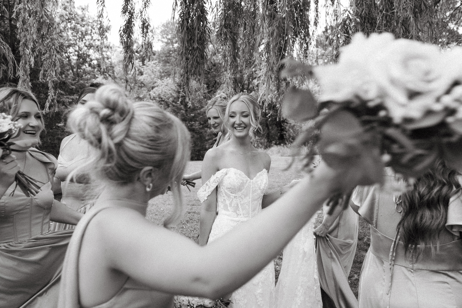Bride smiling with bridesmaids holding bouquets under willow tree in black and white photo.