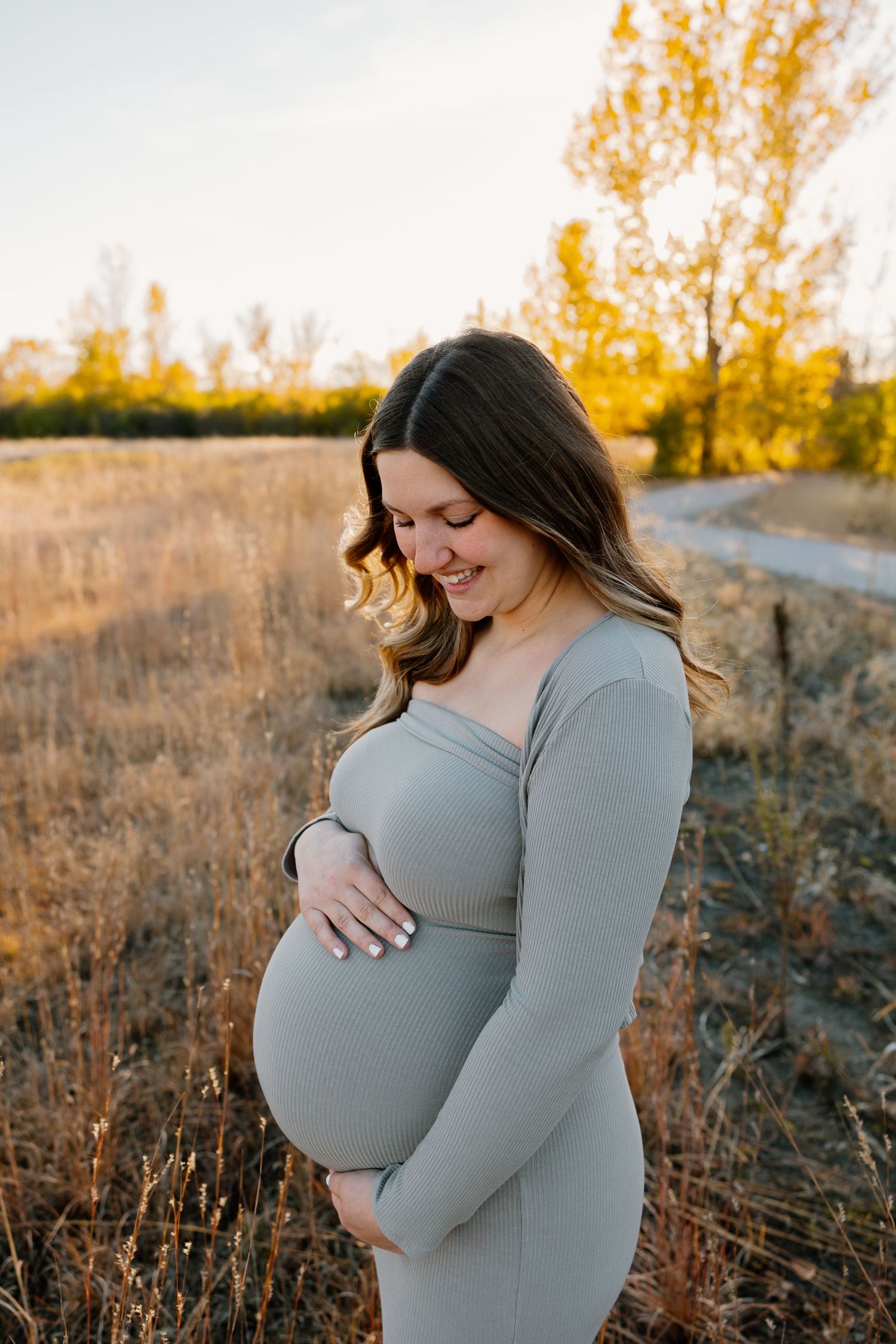 Expectant mother in gray dress cradles baby bump while standing in autumn field at golden hour. Riley Ro Photo Sioux Falls, SD