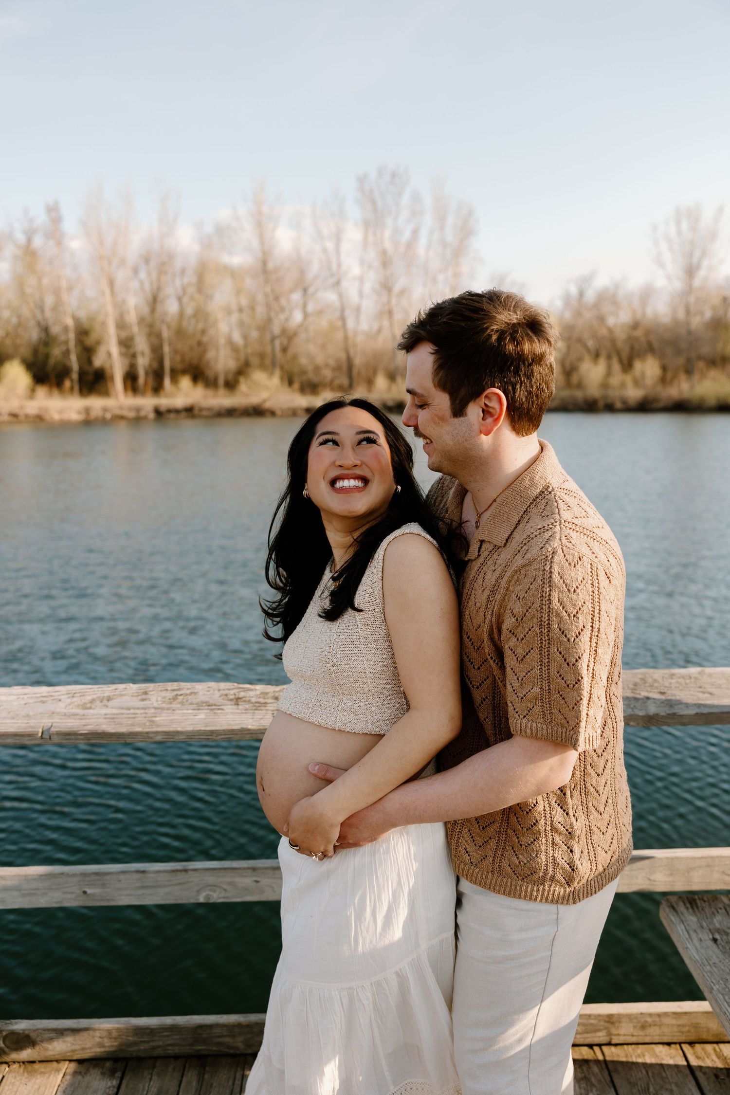 Expecting couple embraces on wooden dock by lake during sunset photoshoot.