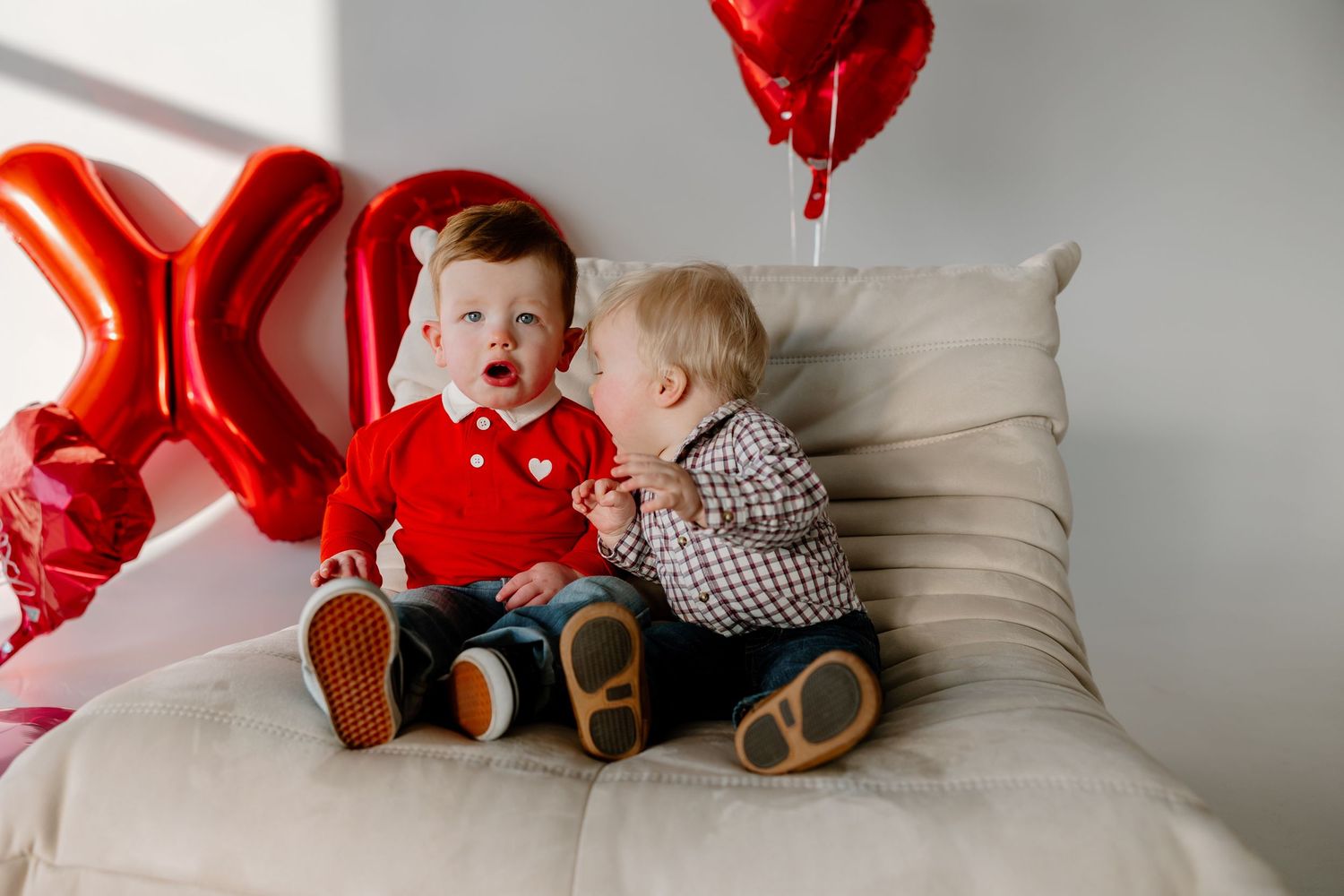 Two children sit on a white couch with red heart balloons for Valentine's Day.