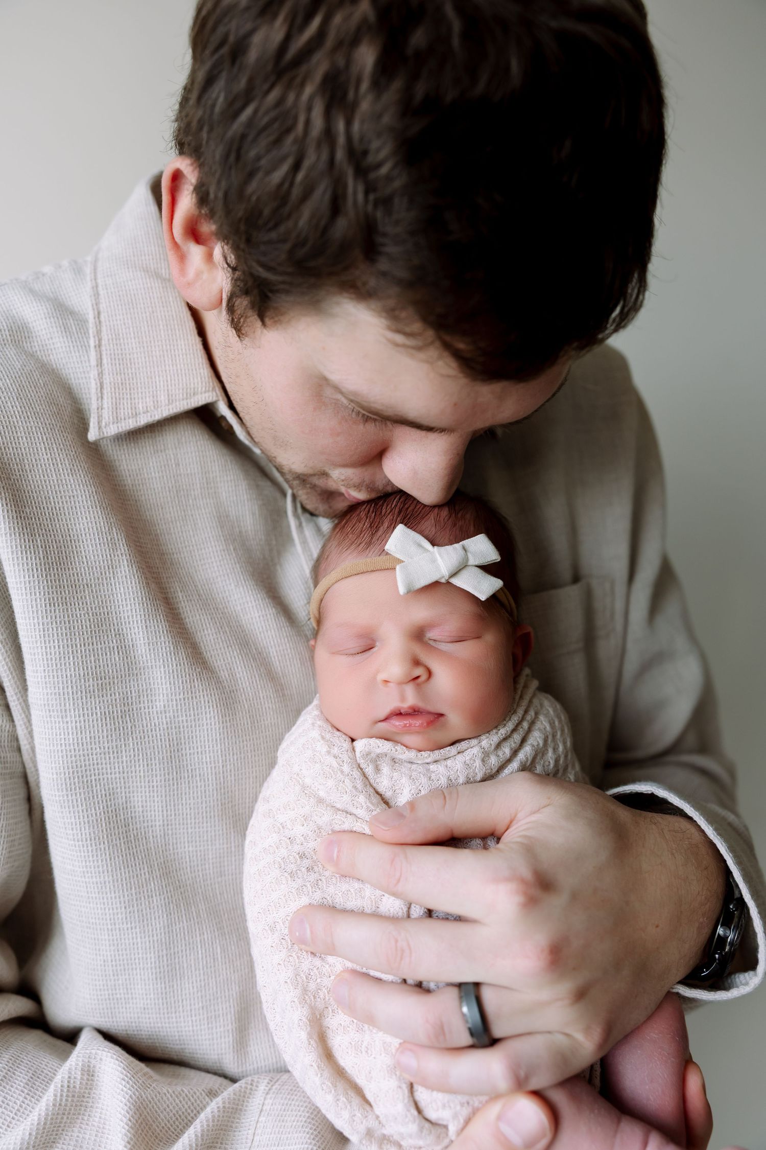 A tender moment between a parent in a beige shirt gently holding and kissing a sleeping newborn in a white bow and swaddle.
