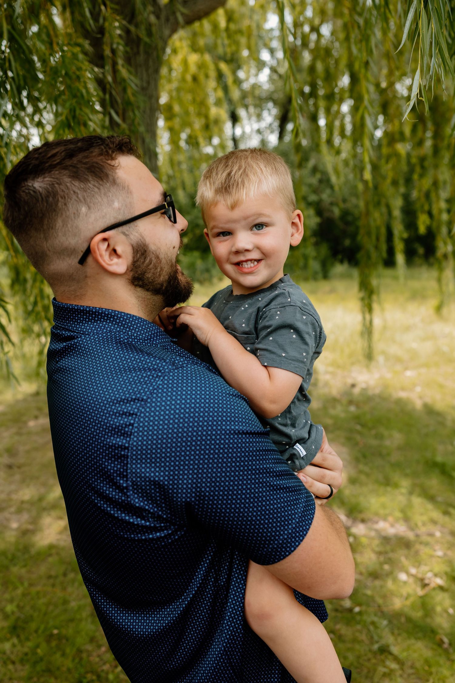 A sweet candid moment shared between a parent in a navy polka dot shirt and a child outdoors under willow trees.