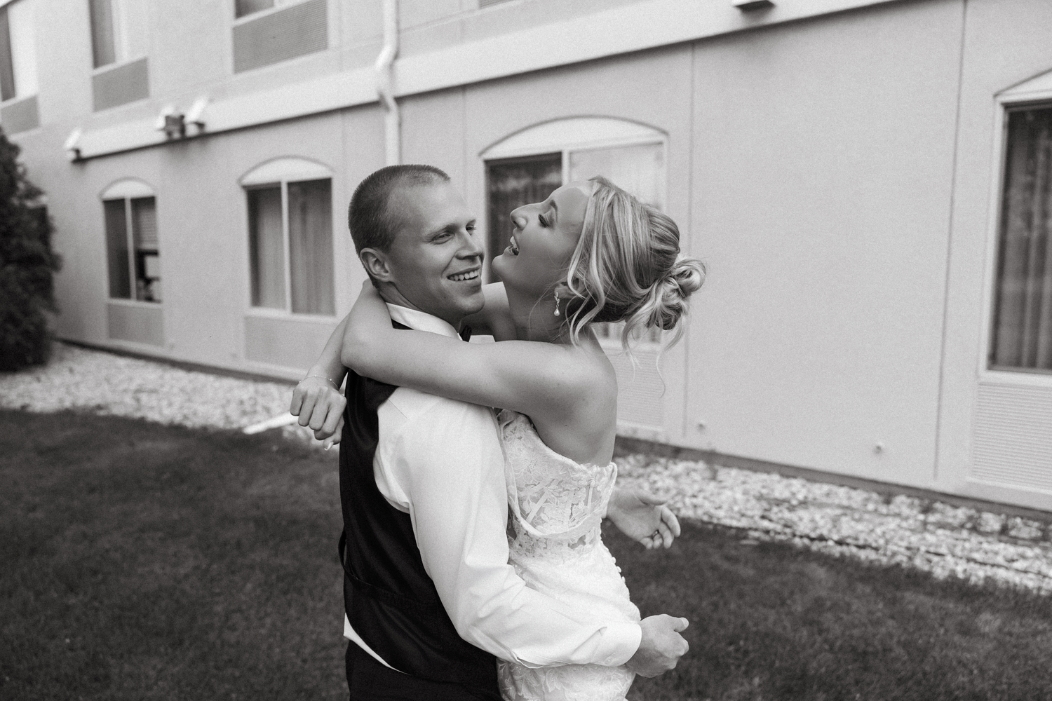 Black and white photo of a groom lifting his bride in a joyful embrace outside a building on their wedding day.