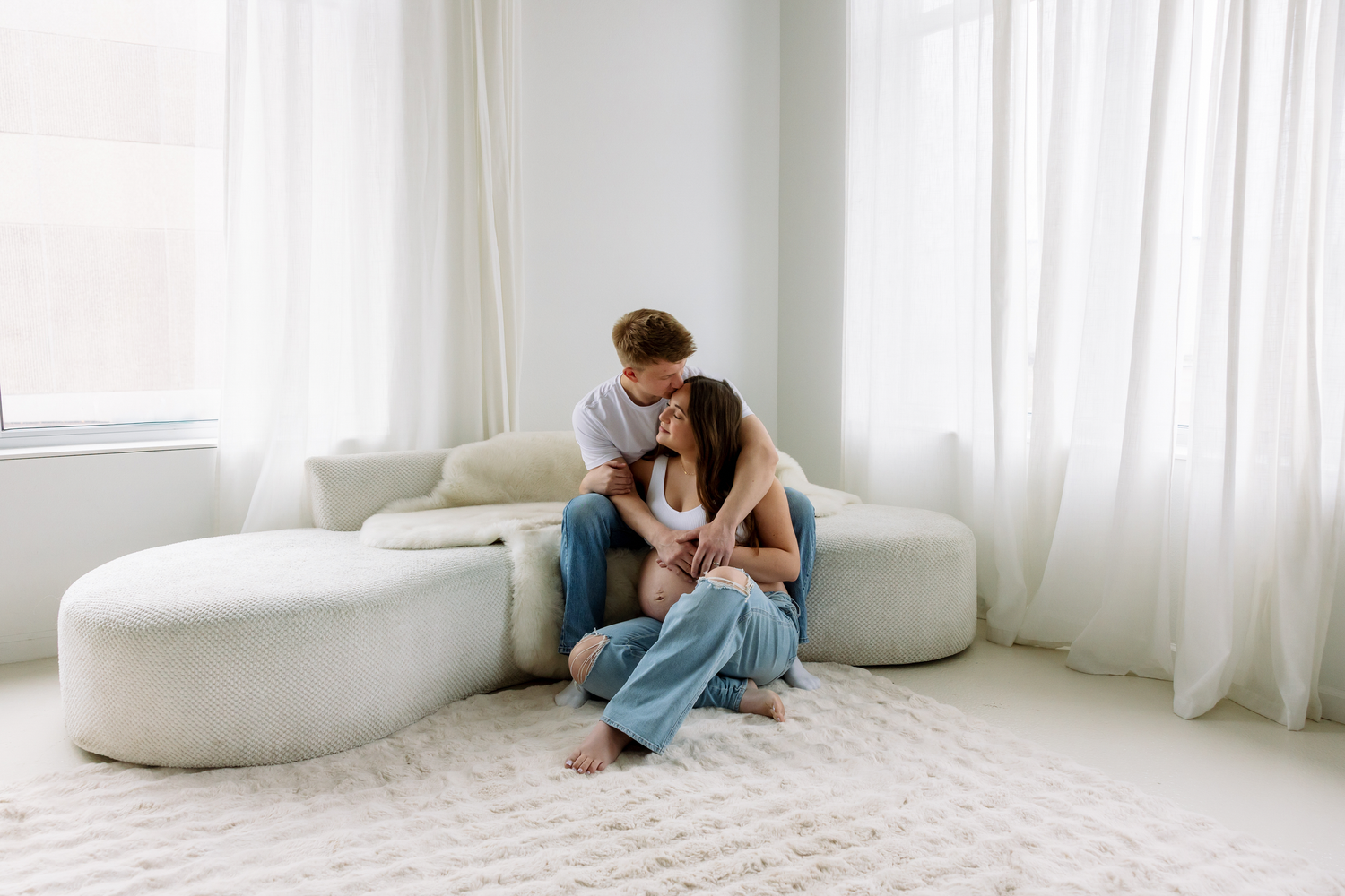 A couple sits on a white rug, sharing a tender moment in a bright, minimalist living room with soft natural light.