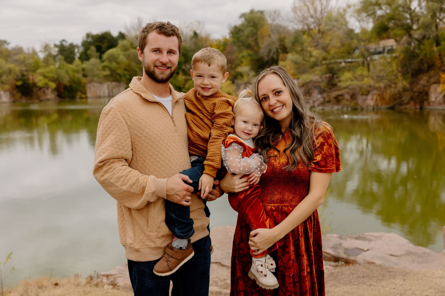 Family of four posing outdoors near a scenic lake, parents holding two young boys, surrounded by autumn foliage.