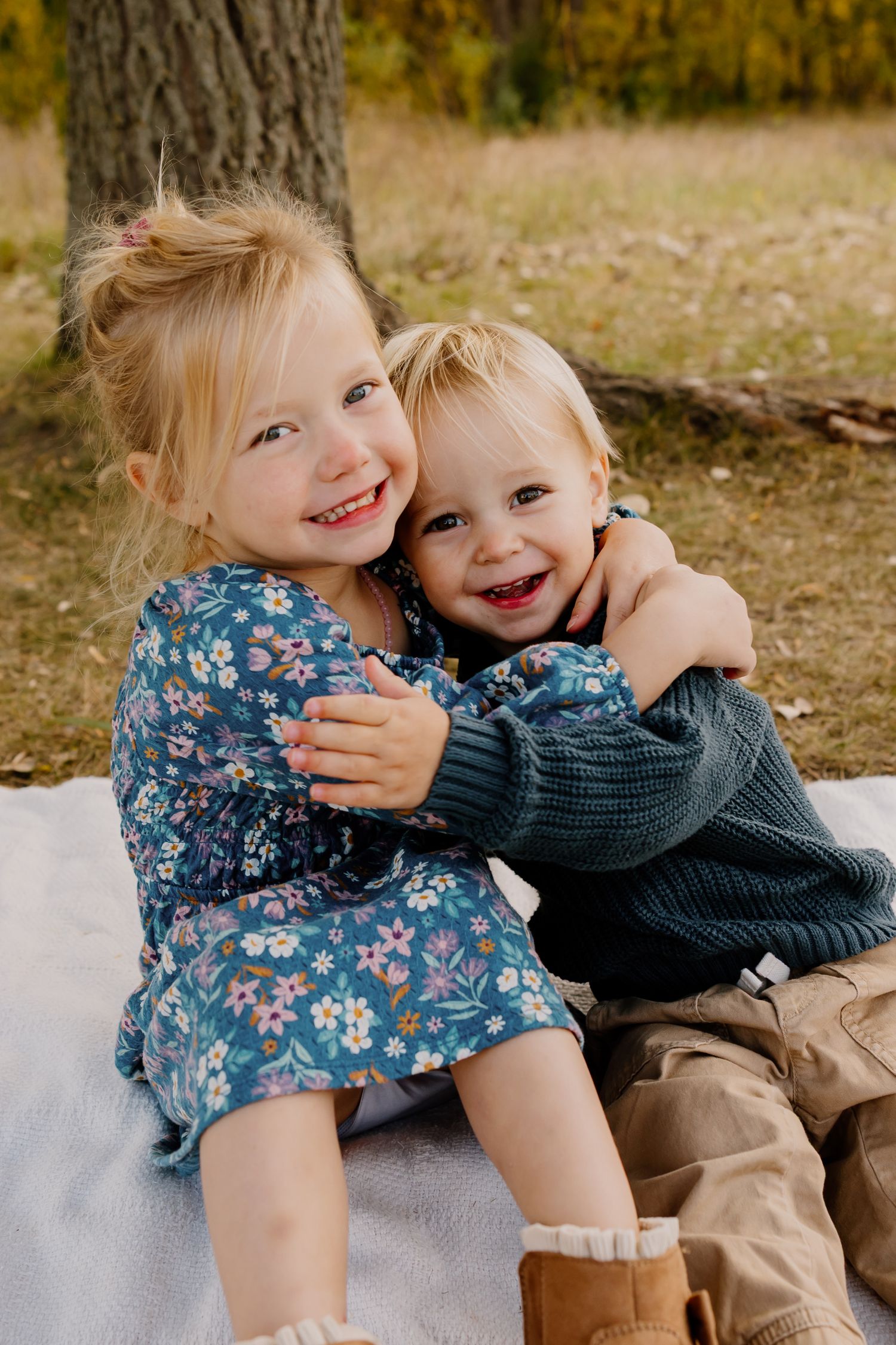 Two young children sitting together outdoors in autumn, wearing coordinated blue and gray outfits and smiling.