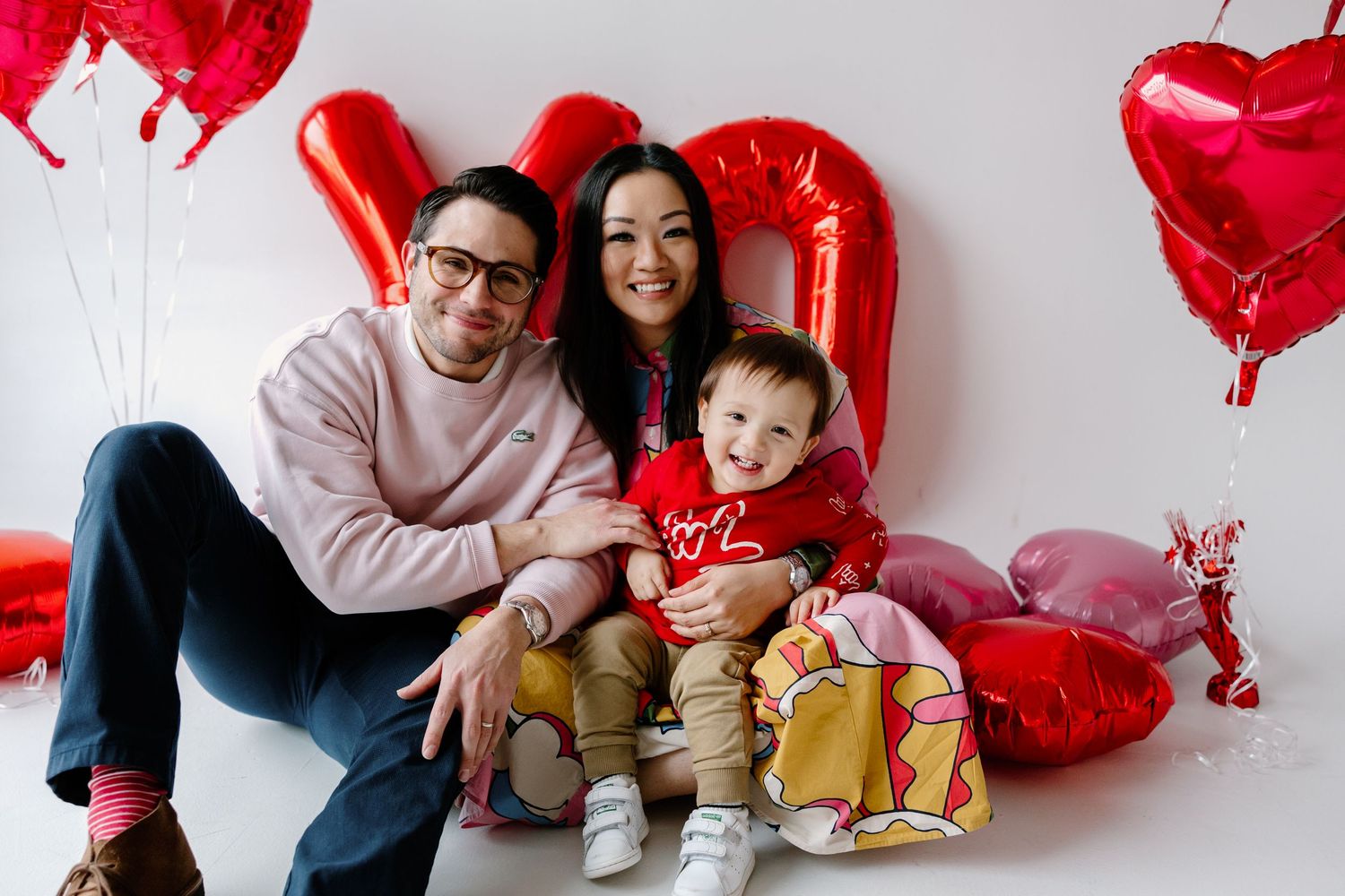 A happy family poses together against a white background with red heart-shaped balloons for Valentine's Day.
