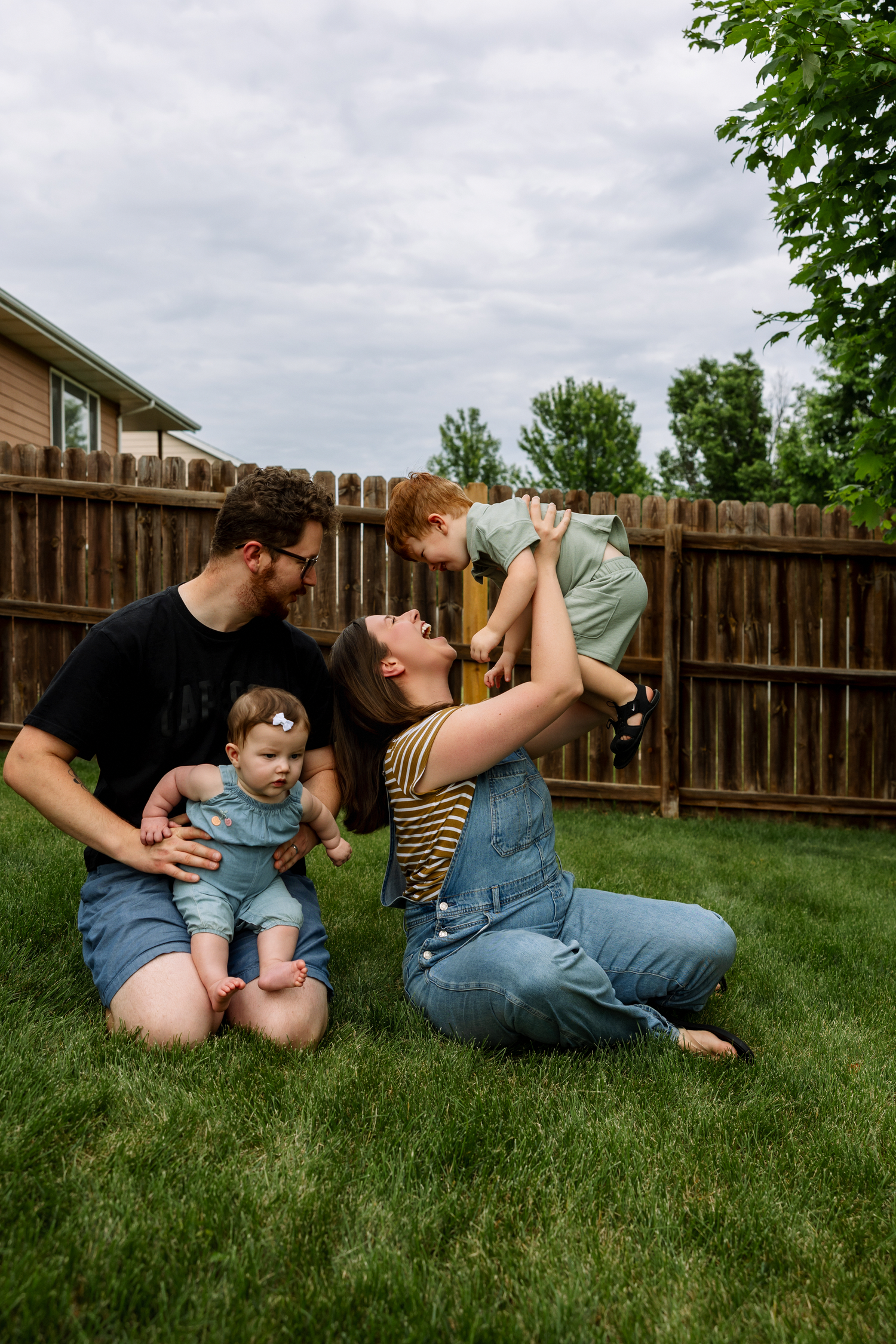 Family of four playing in backyard, parents sitting on grass with baby girl and lifting toddler boy in air.