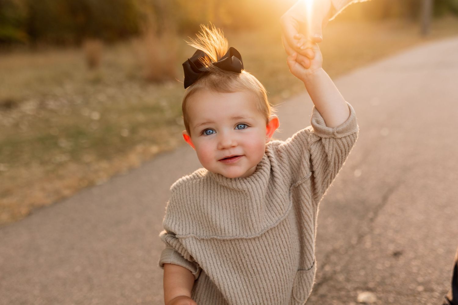 Smiling baby girl with blue eyes and a bun hairstyle raising her arm in golden sunset light outdoors.