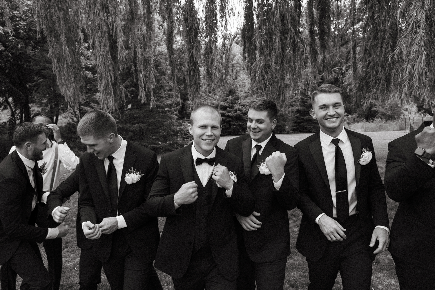Groomsmen in black tuxedos laughing and celebrating outdoors under weeping willow trees at a wedding.