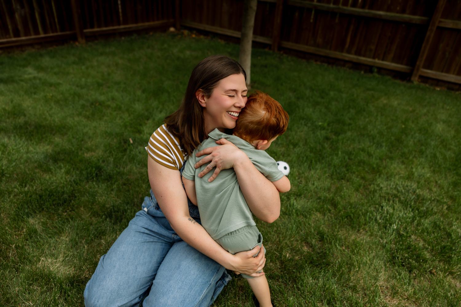 A woman smiling and hugging a red-haired child in a lush green backyard near a wooden fence.