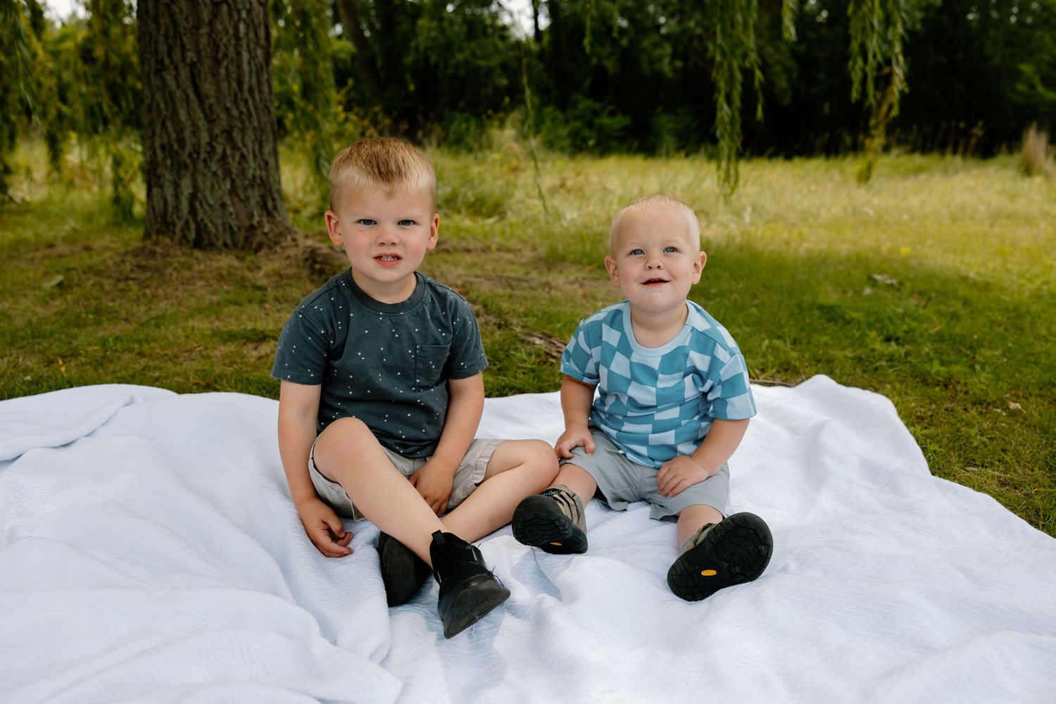 Two small children sitting together on white blanket outdoors with trees in background.