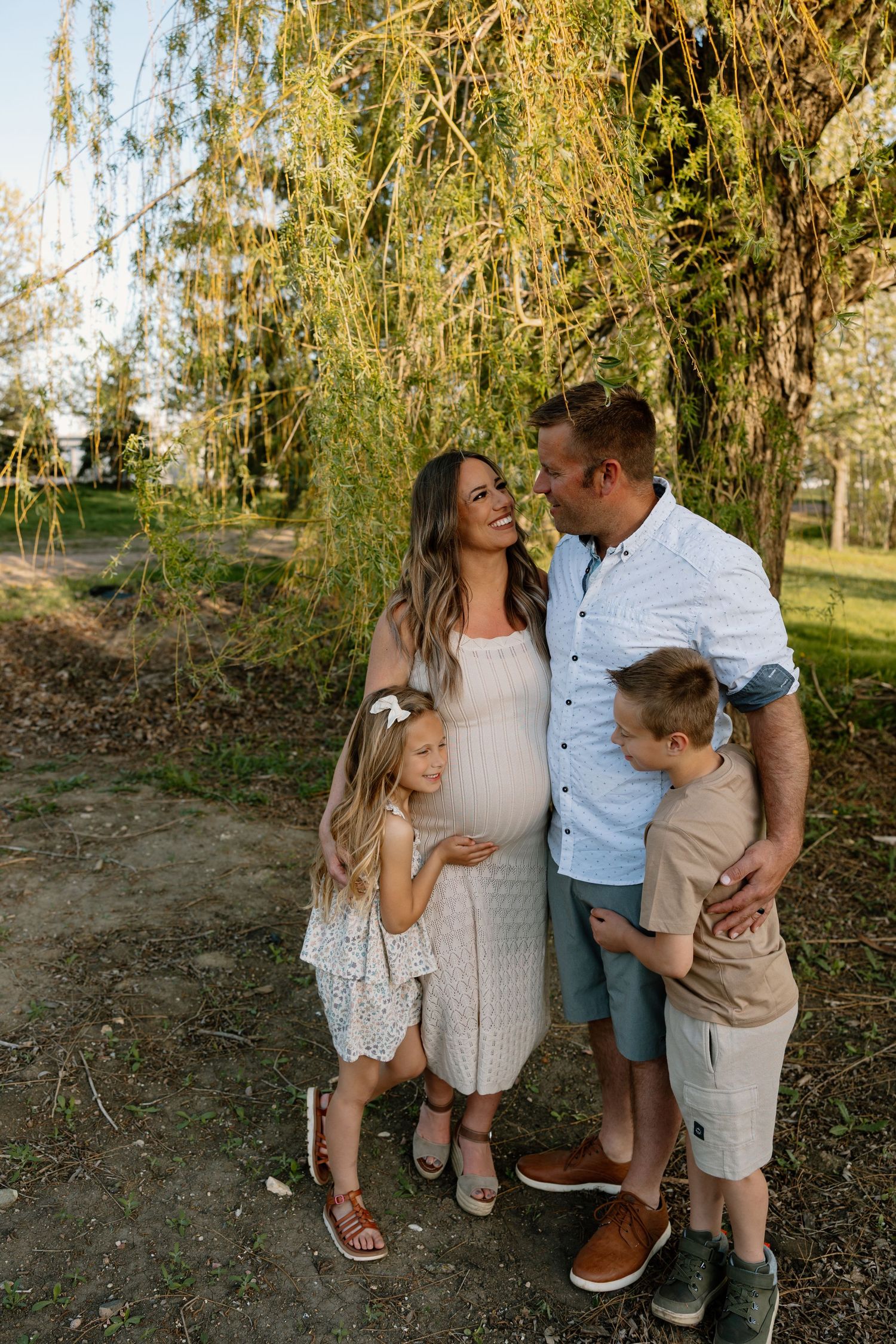 A family shares a tender moment under weeping willow trees in a park during golden hour.