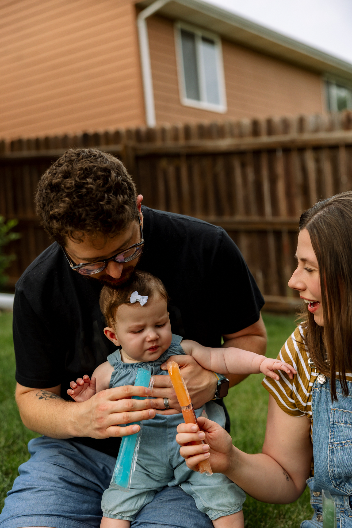 Parents helping curious baby taste a colorful popsicle while sitting together on backyard lawn.