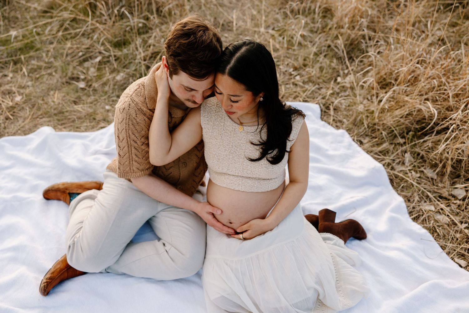 Couple sits intimately on white blanket in golden field sharing tender maternity moment.