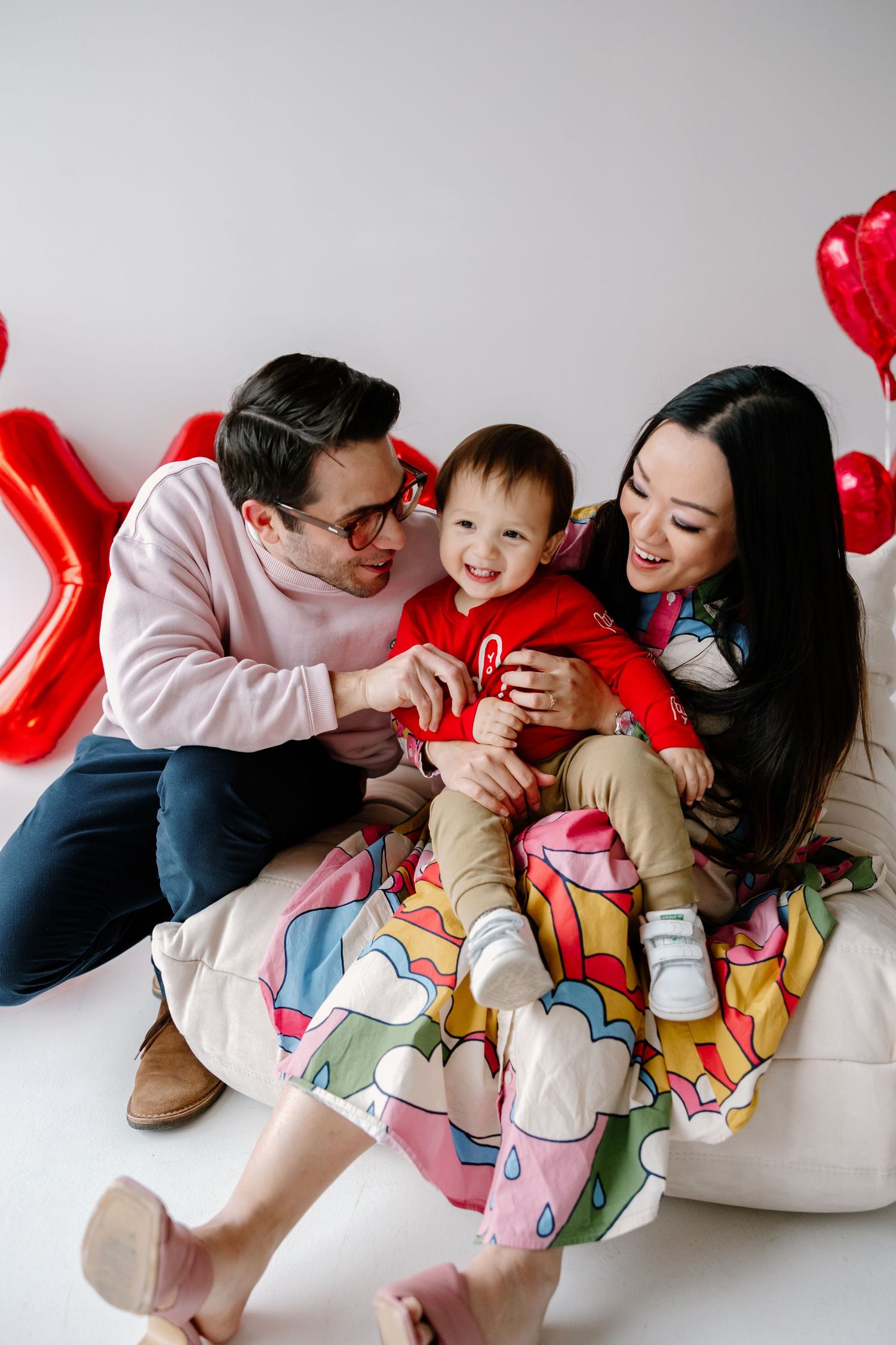 Family sits together on white couch with festive red heart balloons decorating the background for Valentine's Day celebration.