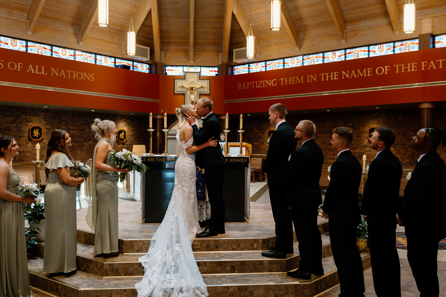 Bride and groom share first kiss at church altar surrounded by wedding party during romantic ceremony.