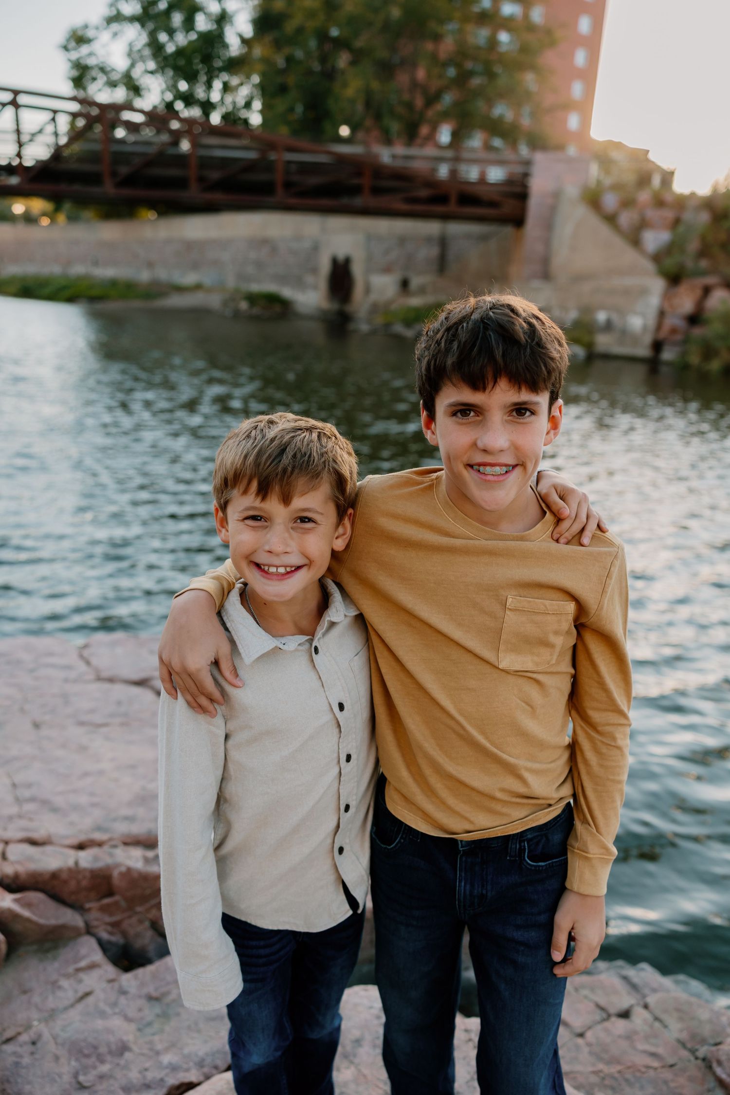 Two children pose together by a waterfront on a sunny day, wearing casual clothing and sharing warm smiles. Riley Ro Photo Sioux Falls, SD