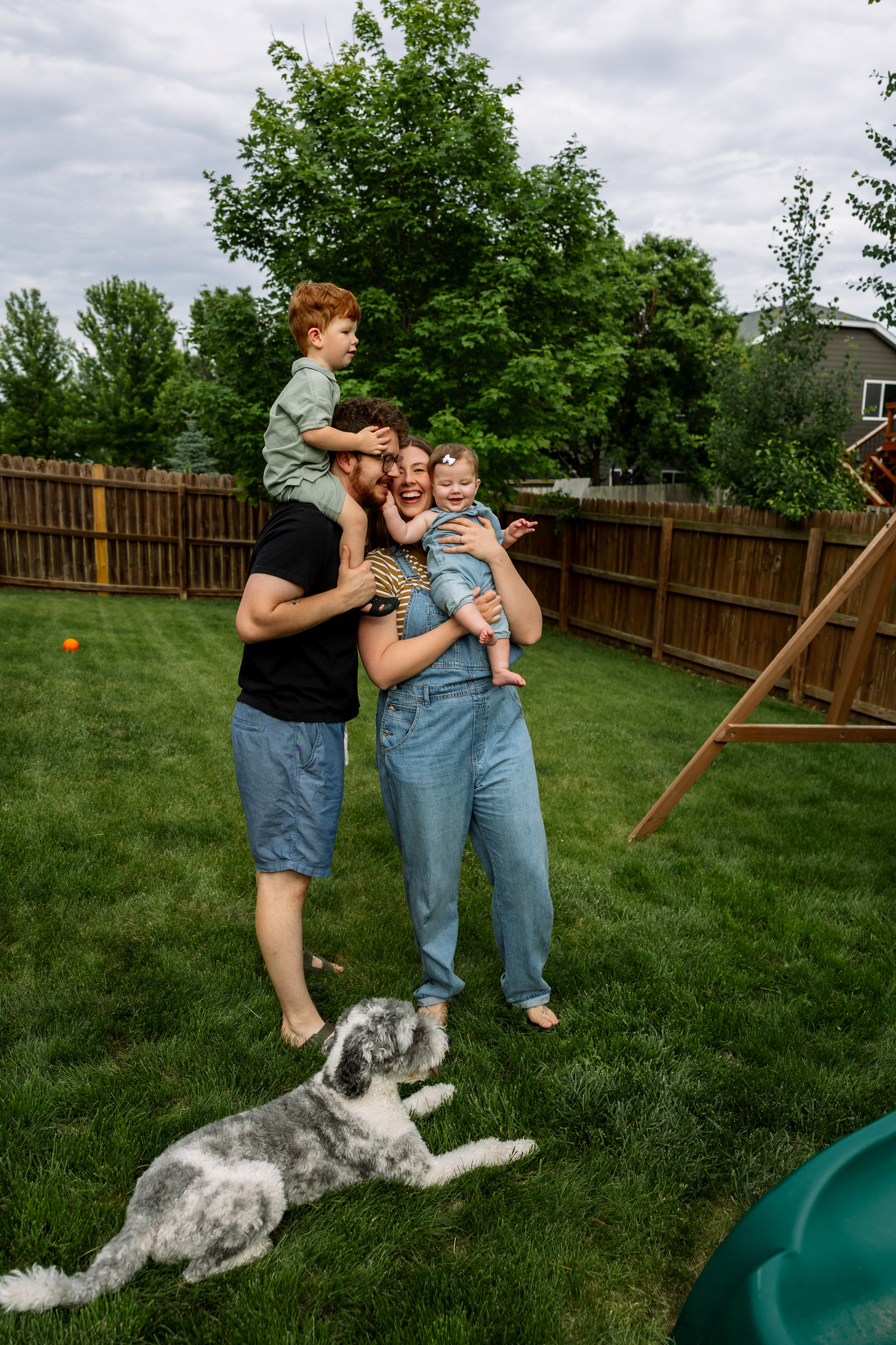 Family with two kids and a fluffy dog enjoying a sunny day in a fenced backyard with green grass.