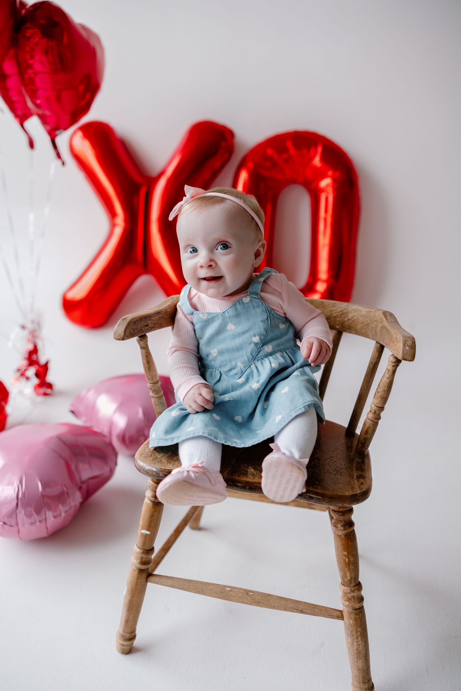 A baby in light blue attire sits on wooden chair with red and pink heart shaped balloons in background.