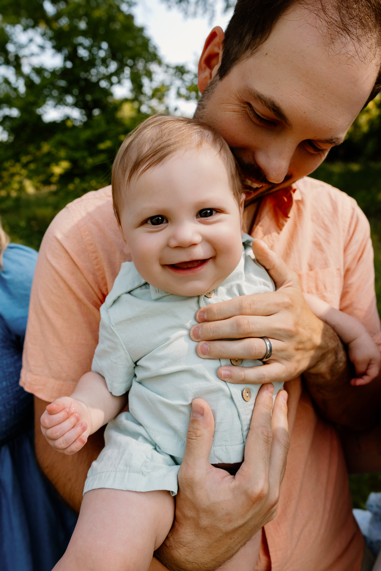 A smiling baby boy held by his father outdoors during a family photo session in a lush green park.