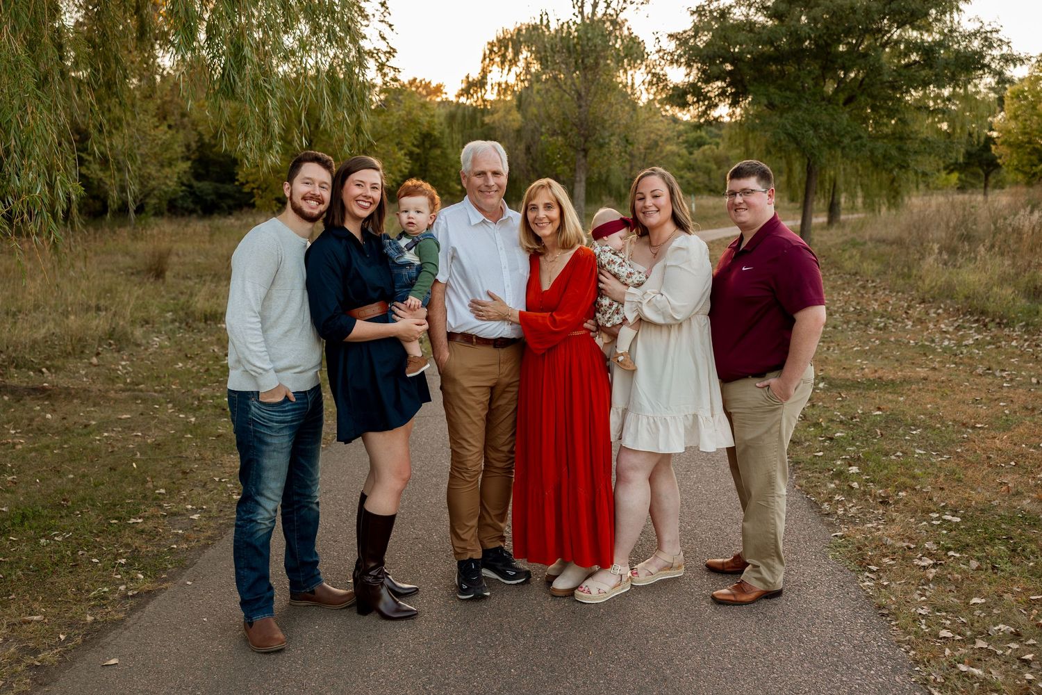Family of seven smiling together outdoors on a path surrounded by green trees during golden hour.