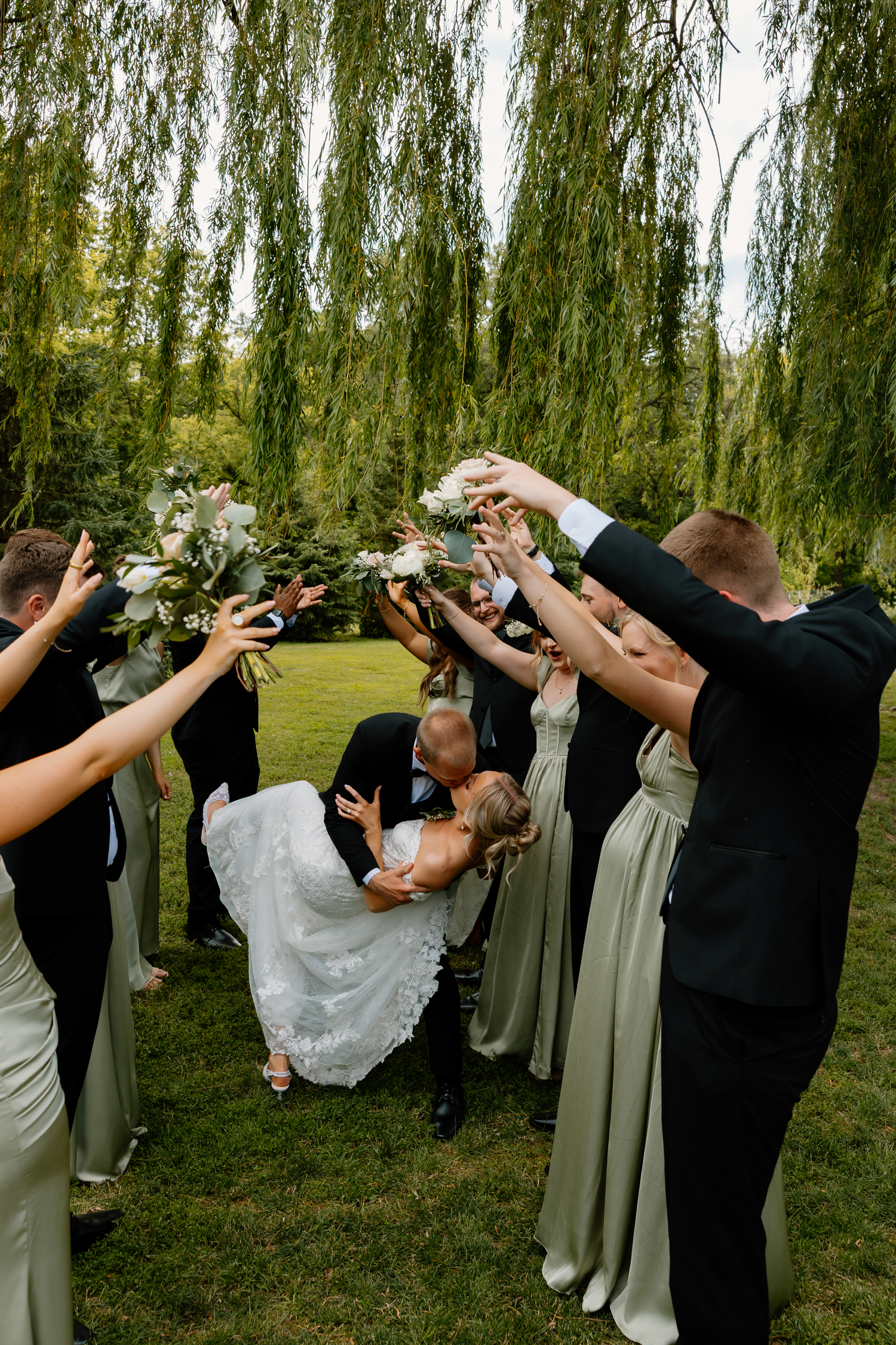 Wedding party cheering as bride and groom share a romantic dip kiss under a weeping willow tree outdoors.