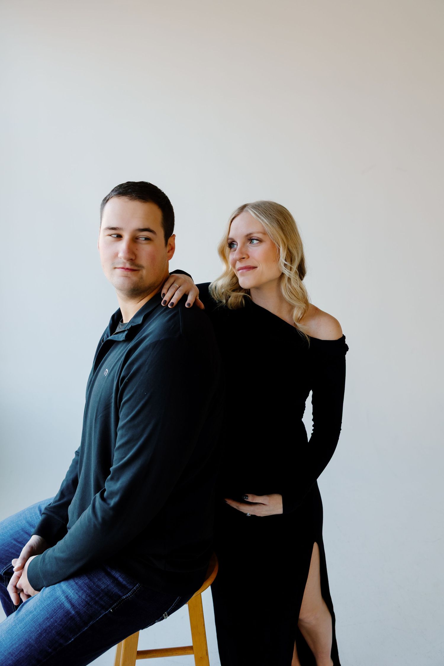 A stylish maternity photo featuring a couple in dark clothing sitting on a wooden stool. Riley Ro Photo Sioux Falls, SD
