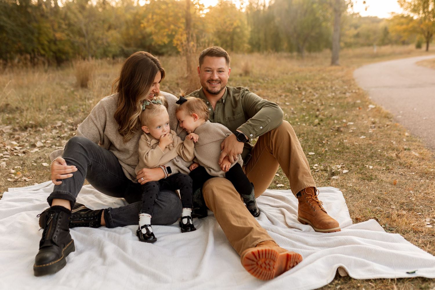 Family of four sitting on white blanket outdoors in autumn park, parents smiling with two young children.