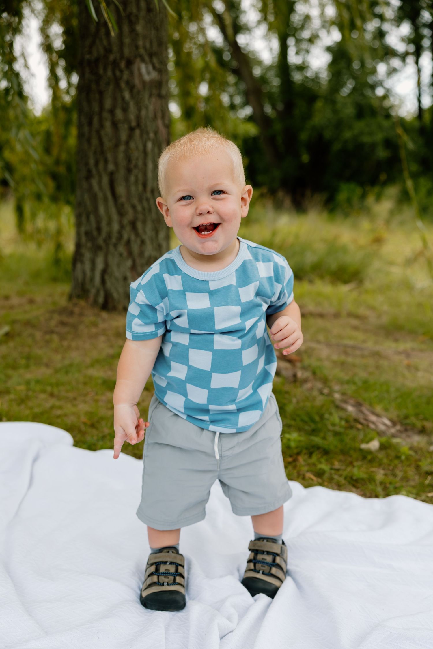 Happy toddler wearing blue checkered shirt and gray shorts stands on white blanket outdoors.