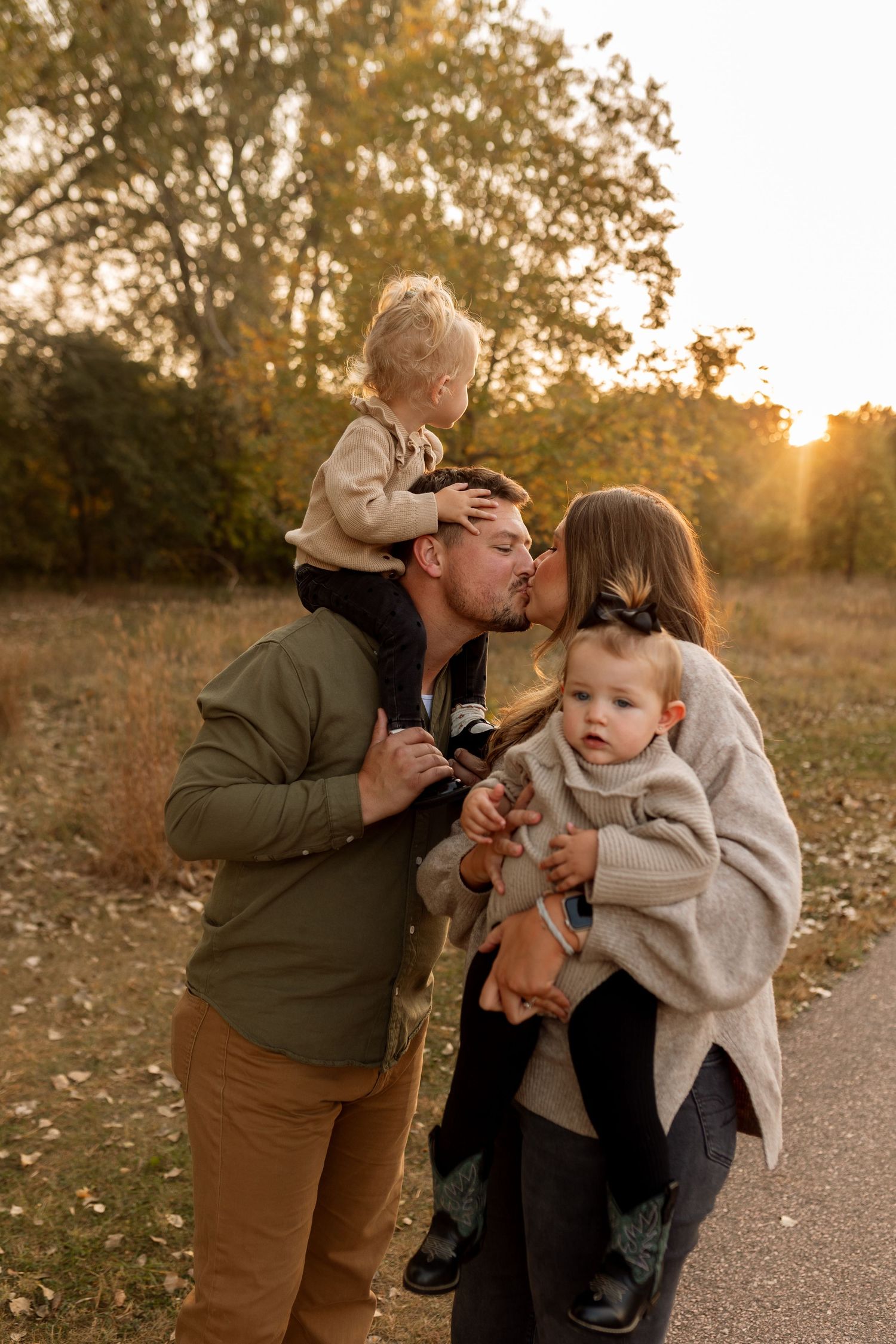 A young family shares a kiss at golden hour, with dad holding a toddler on shoulders and mom cradling a baby outdoors.