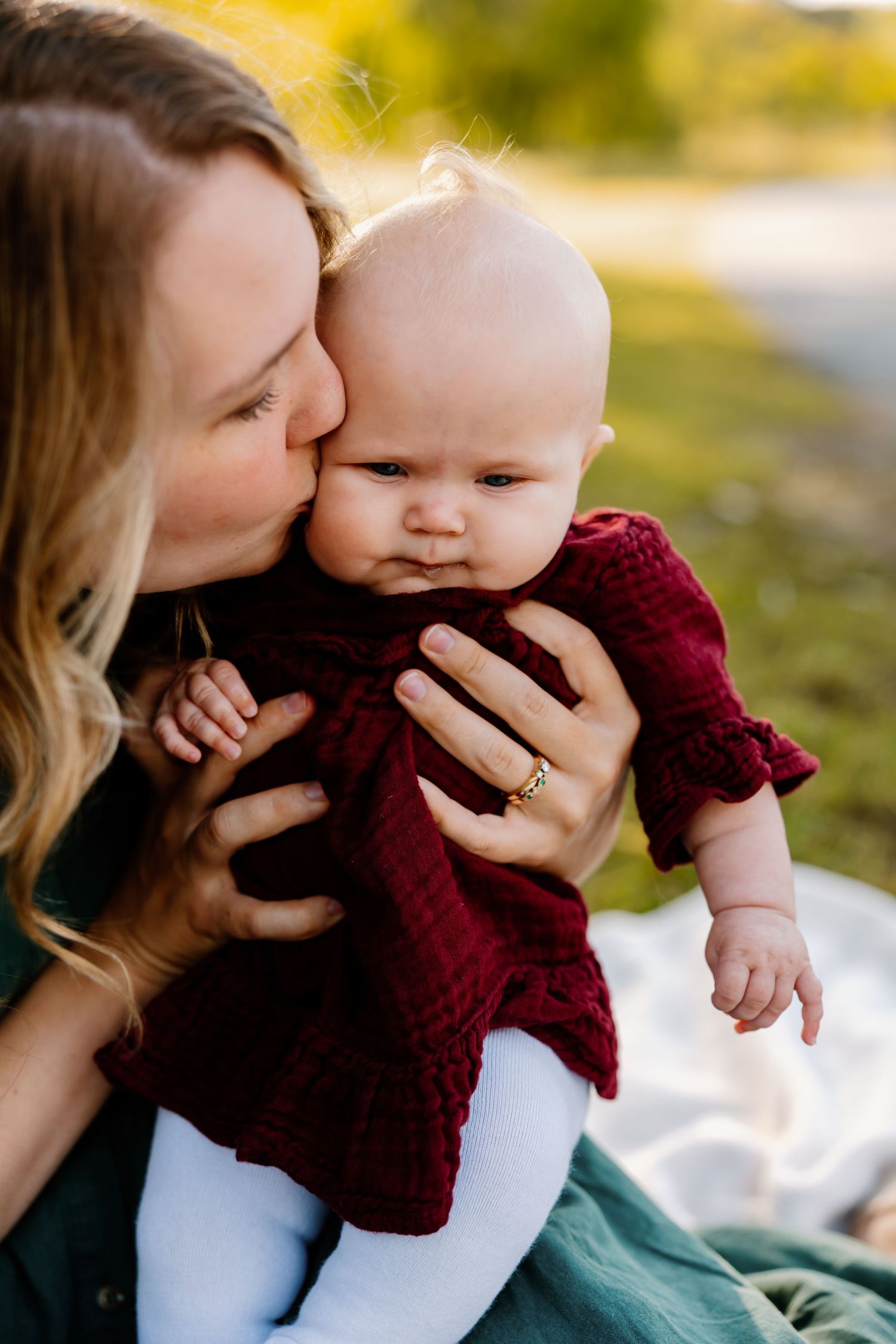 A mother tenderly kisses her baby girl in a red dress outdoors, capturing a warm and loving moment.