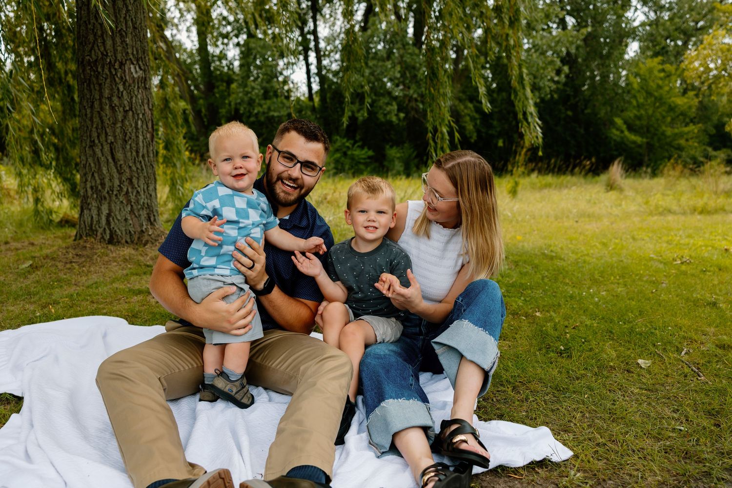 Family sitting together on white blanket for outdoor photo session in natural grassy setting during golden hour.