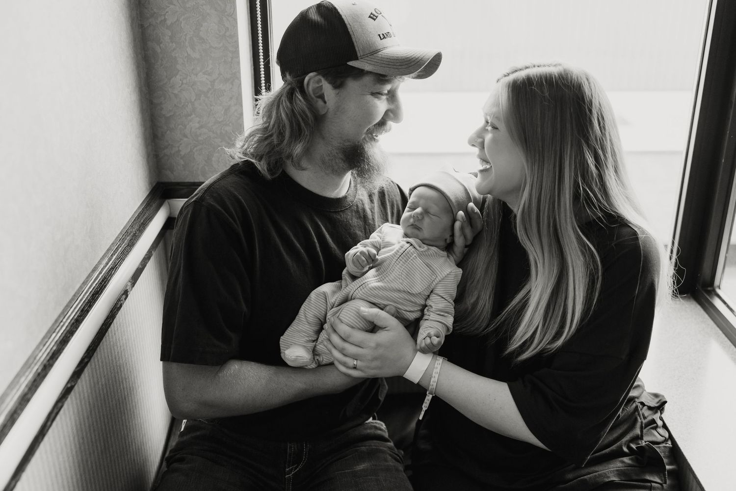 New parents share a tender moment while holding their newborn by a window.