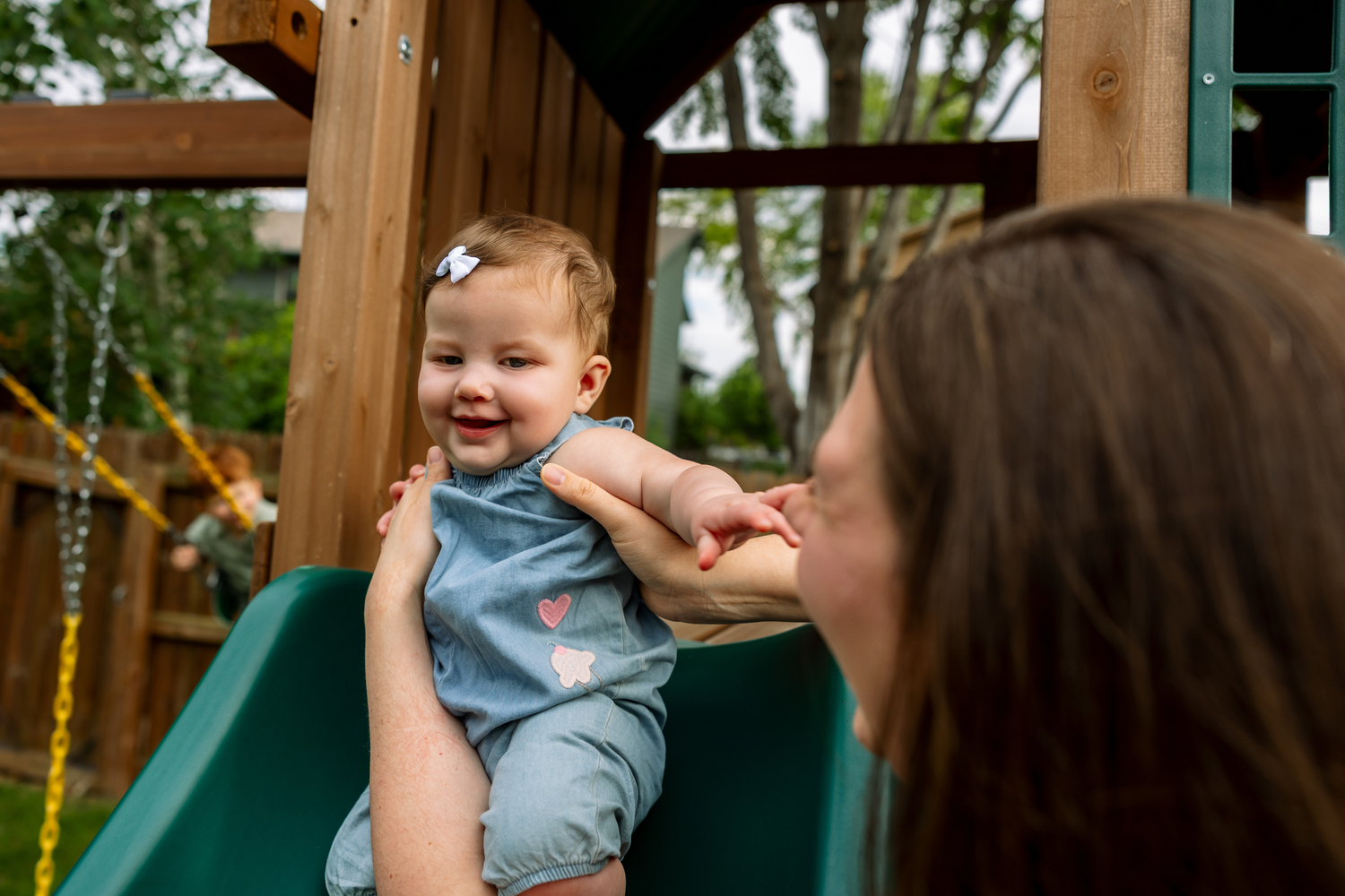 A mother helps her smiling baby girl on a green slide at a wooden backyard playset.