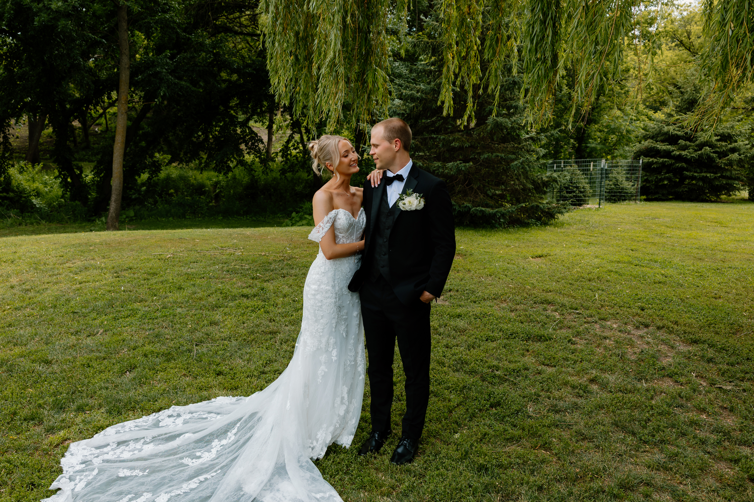 Bride and groom share a romantic moment on green lawn beneath a weeping willow tree in wedding attire.