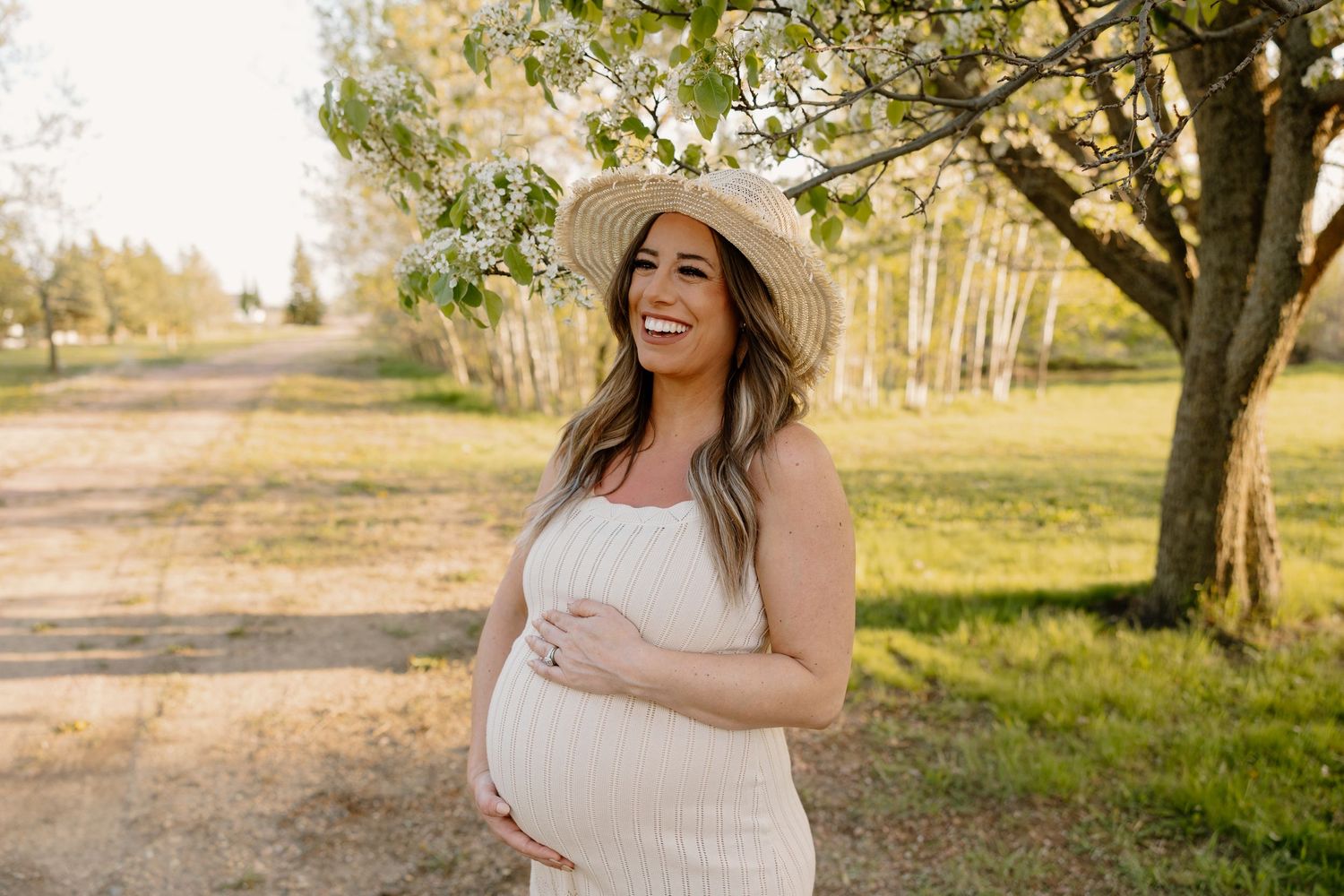 Person in white dress and straw hat stands joyfully on a dirt path surrounded by trees during golden hour.
