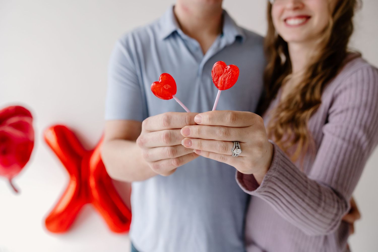Close up of hands holding heart-shaped lollipops with red balloon decorations in the background.