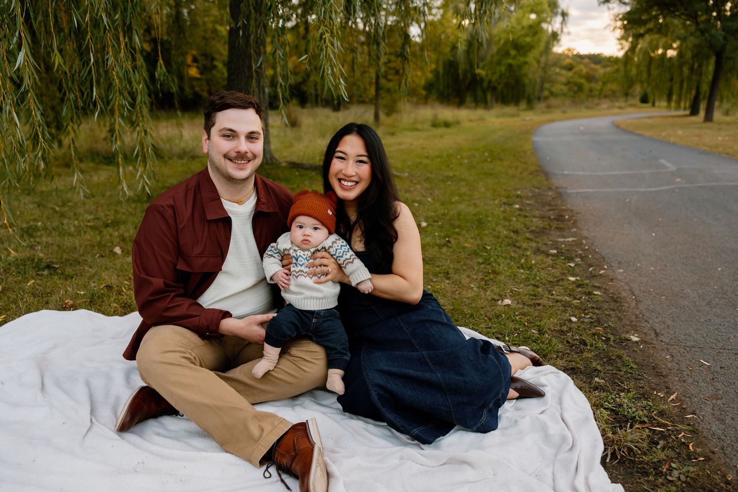 A family sits together on a white blanket during an autumn outdoor photoshoot along a tree-lined path. Riley Ro Photo Sioux Falls, SD