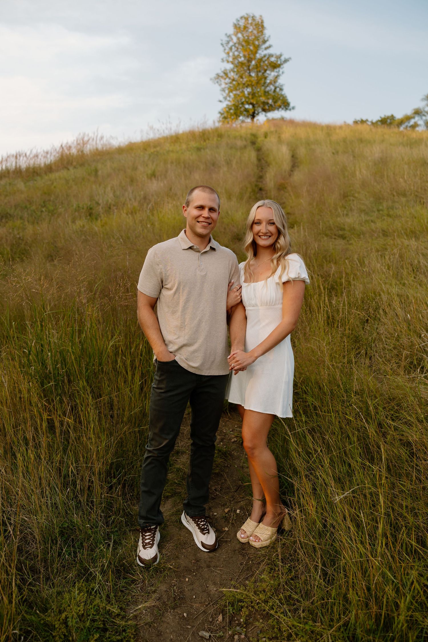 A couple standing on a hill path at golden hour with a tree silhouetted against the sky.