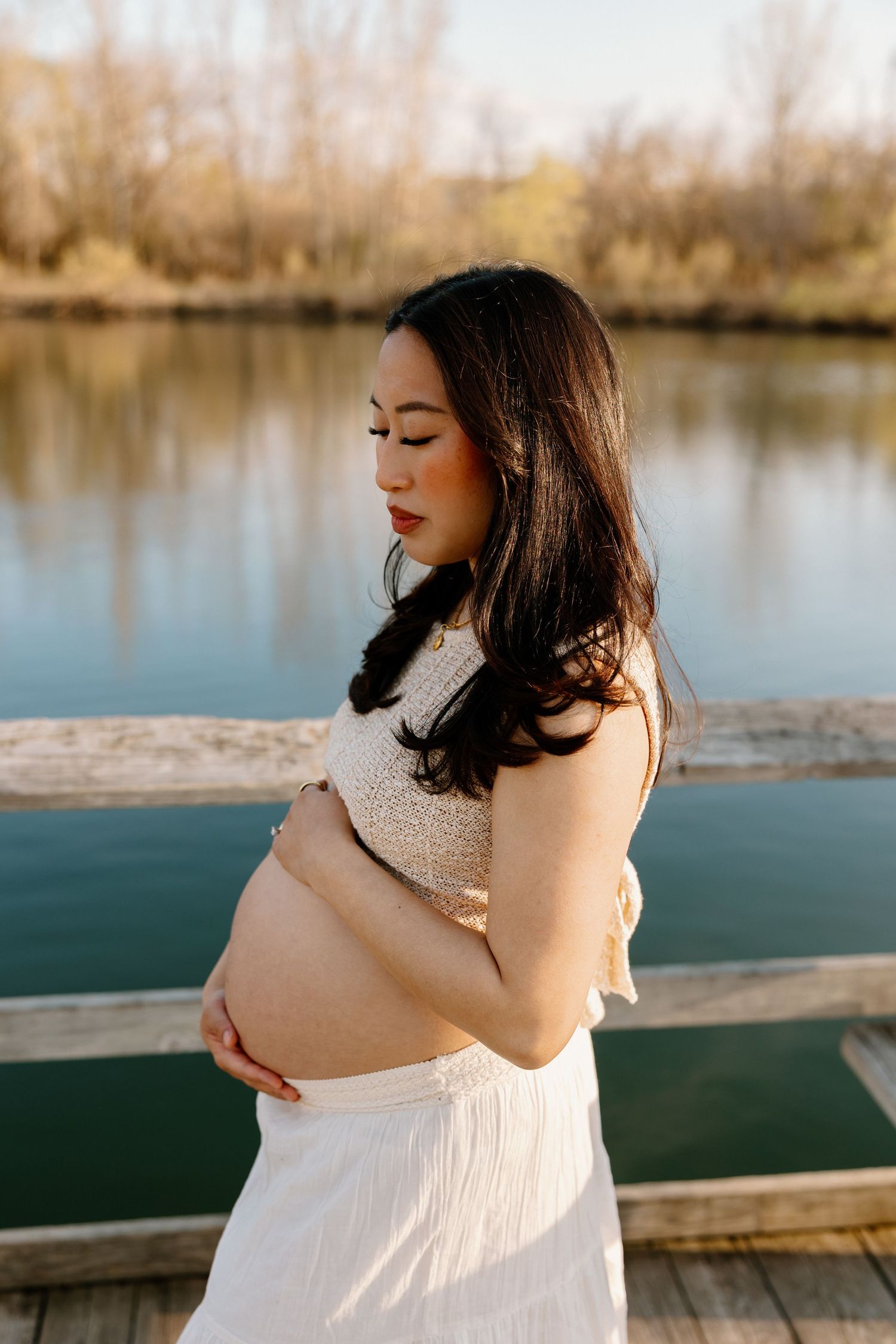 An expectant mother in white dress poses on a dock during golden hour, cradling her baby bump.