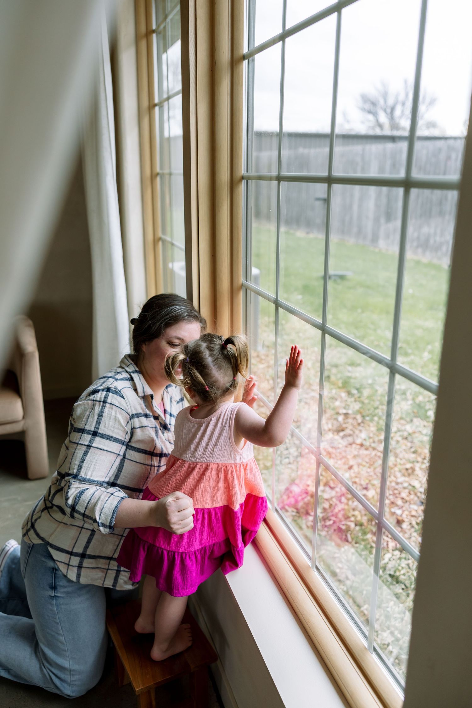 A mother kneels beside her toddler daughter in a pink dress as they look out a large window together.