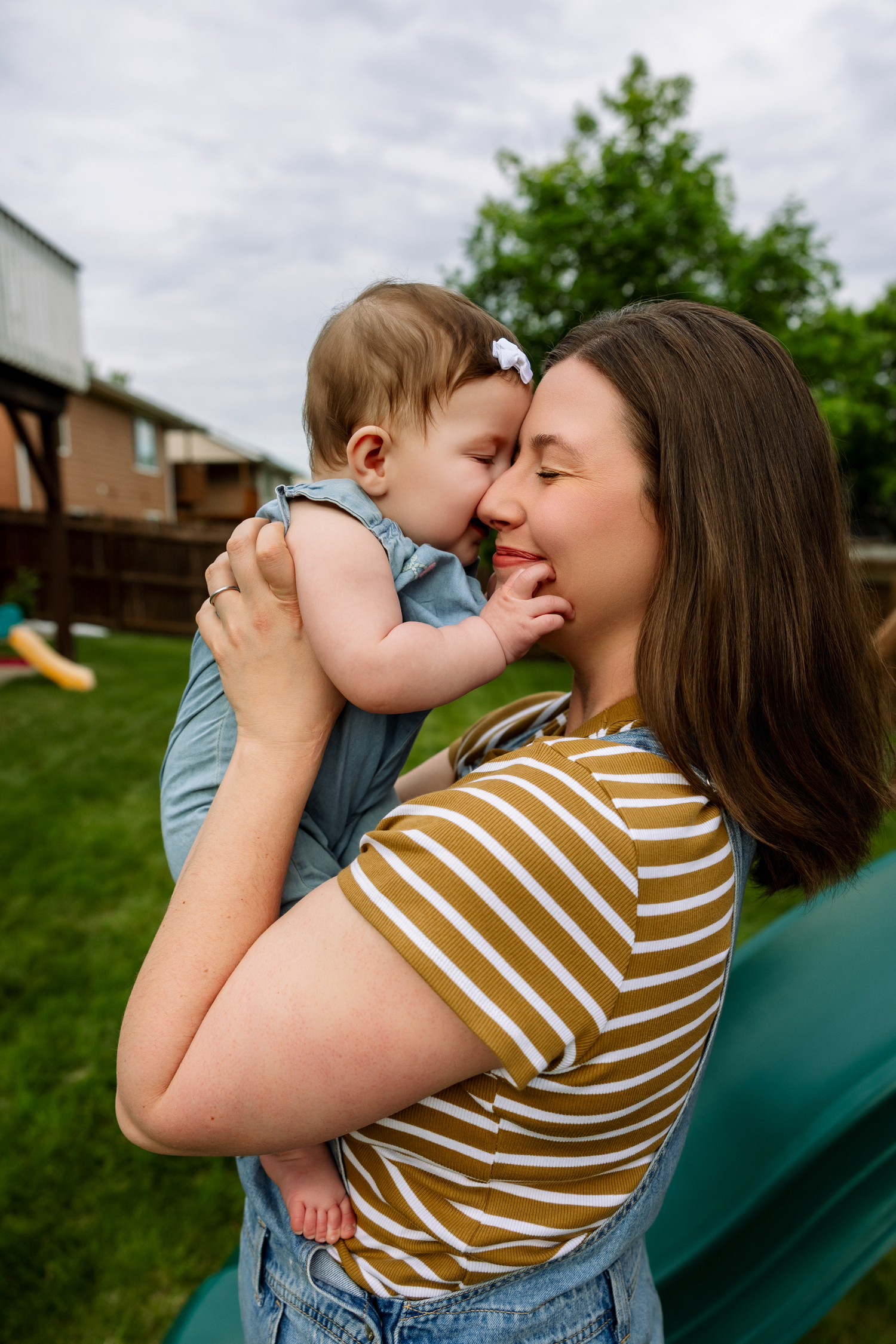 A young mother in a striped shirt lovingly holds her baby girl outdoors in a green backyard.
