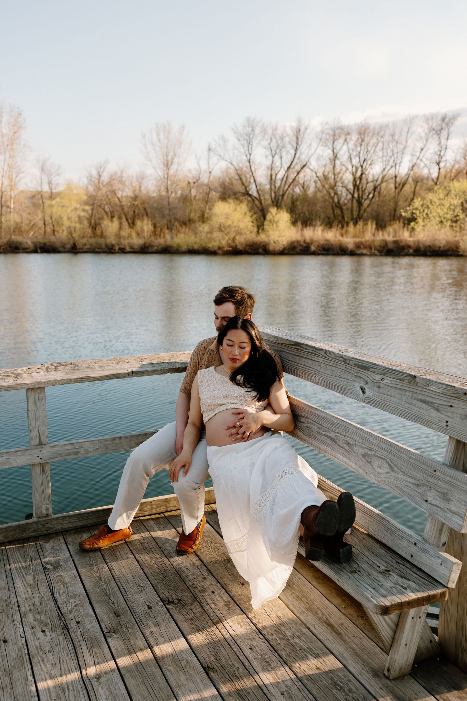 A couple sits together on a wooden dock bench overlooking a serene lake during golden hour.
