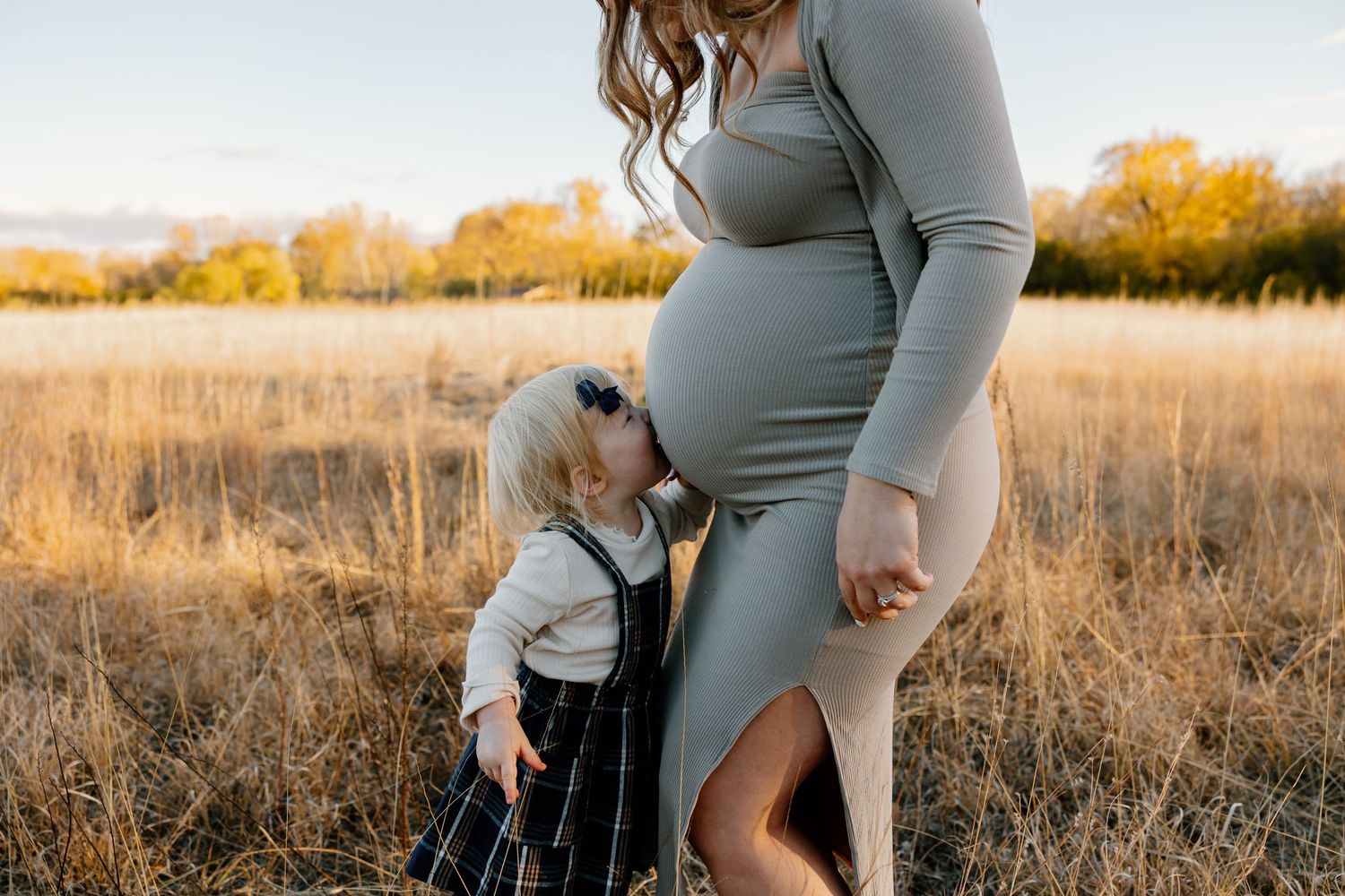 A toddler in a gray outfit touches a pregnant woman's belly while standing in a golden autumn field at sunset.