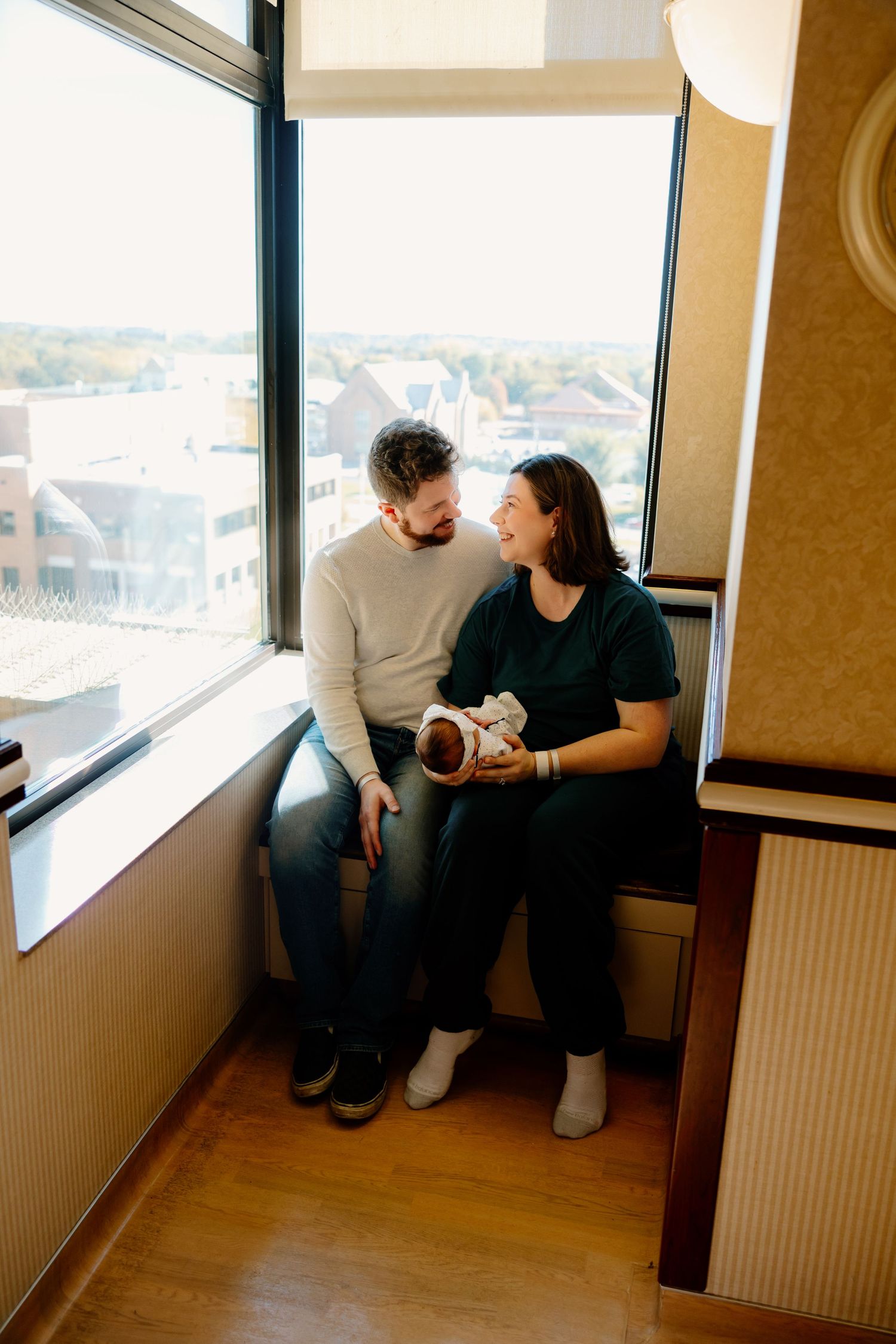 Couple sharing a cozy moment by large window in apartment building with cityscape view in background. Riley Ro Photo Sioux Falls, SD